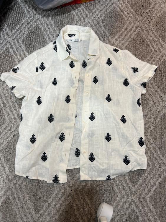 White short-sleeved shirt with black geometric pattern, on a gray rug. Visible tag on the collar.