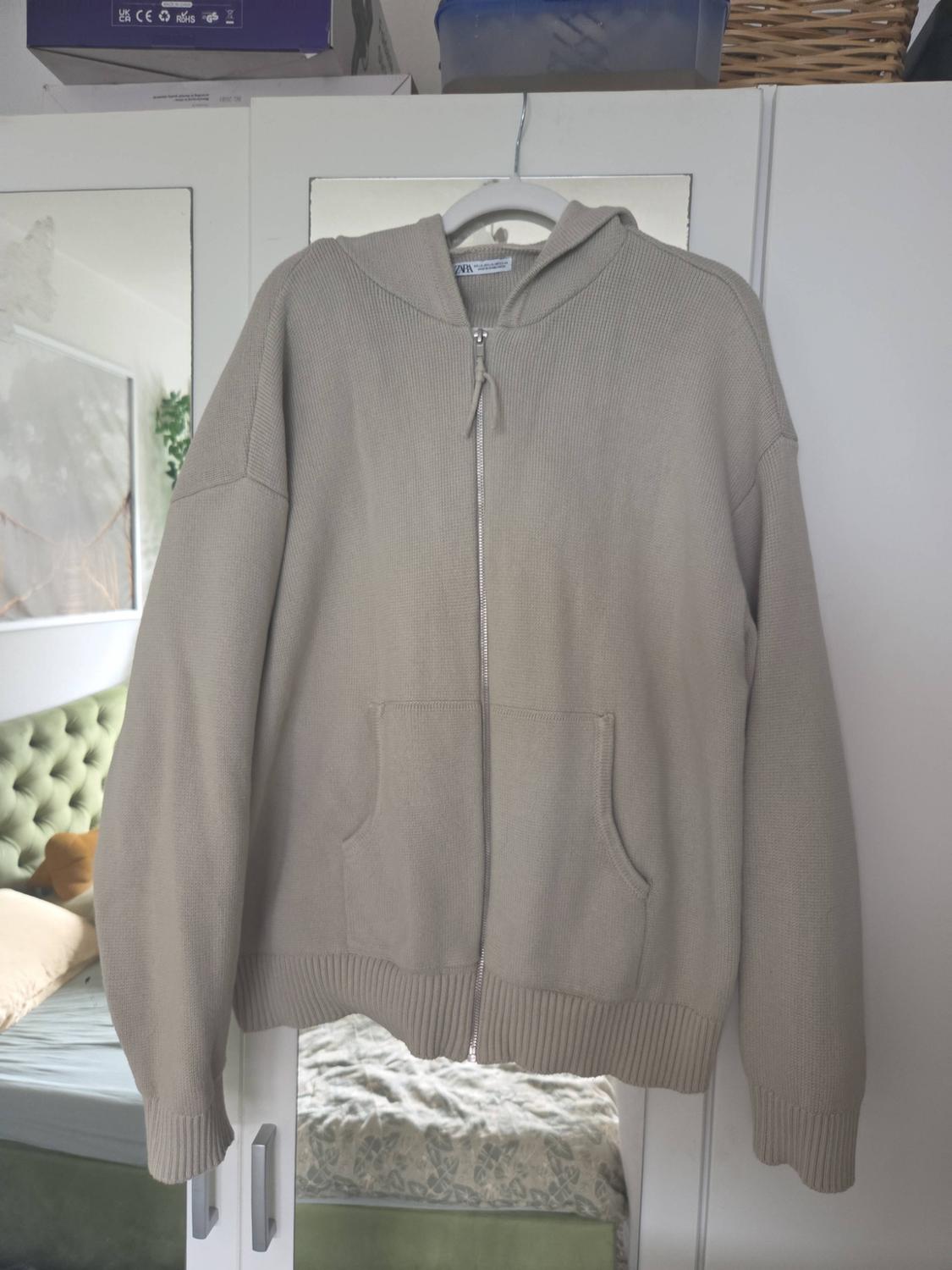 Beige knitted zip-up hoodie with a hood hanging in a wardrobe. The mirror reflects a bed. Texts: ZARA, UK CA CE RoHS GS.