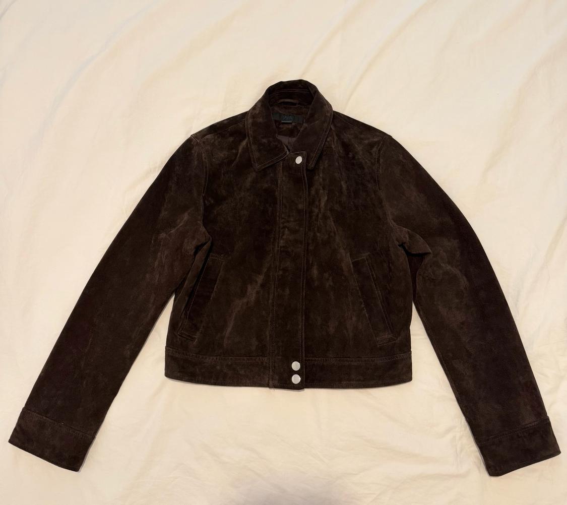 Dark brown suede jacket with a collar and buttons, laid out on a creased light-coloured fabric.