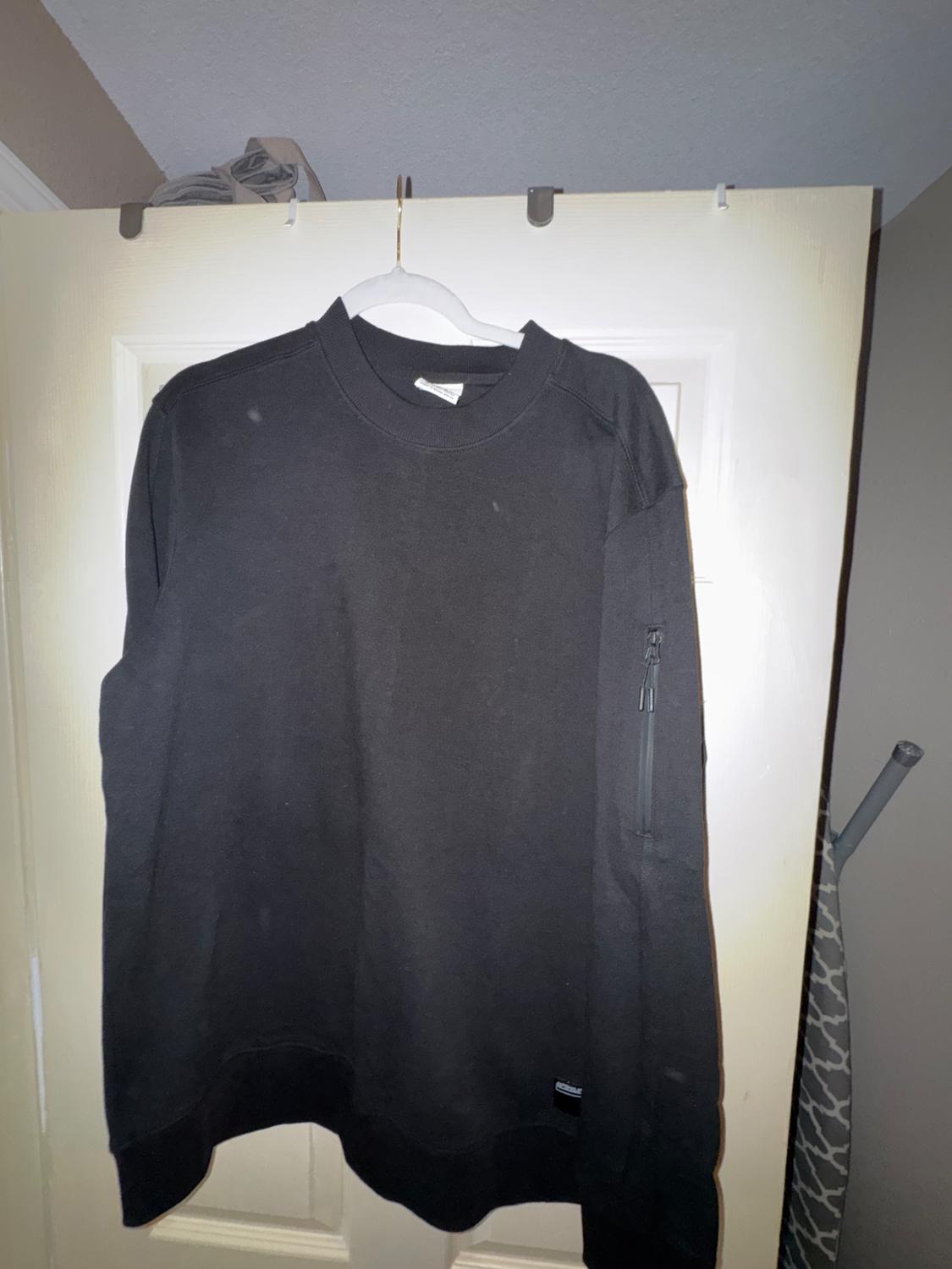 Black sweatshirt hanging on a white hanger over a door. It has a zipped pocket on the left sleeve.