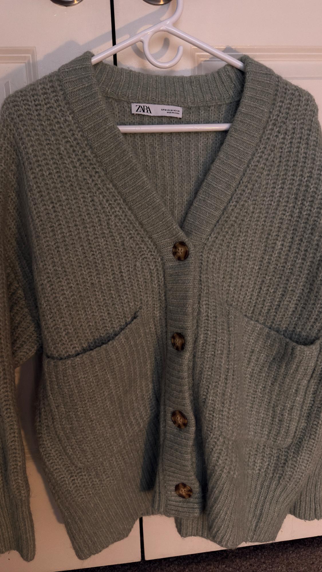 Gray knit cardigan with brown buttons and front pockets, hanging on a white hanger. Visible tag: "ZARA".