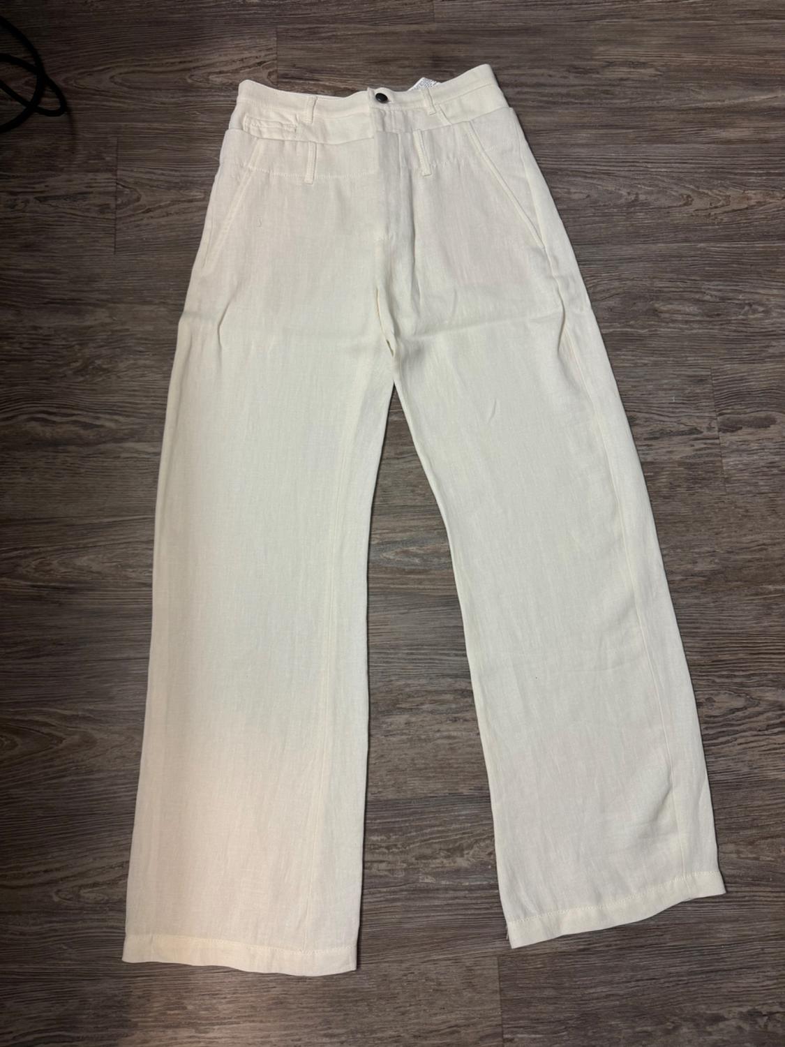White, straight-leg trousers with front pockets, black button and zip fastening, on a brown wooden floor.