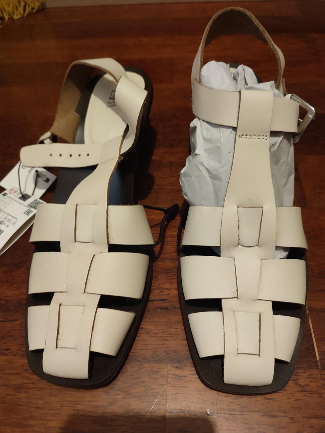 Beige leather sandals with an interwoven design on a wooden floor. One sandal has paper inside and a visible label.