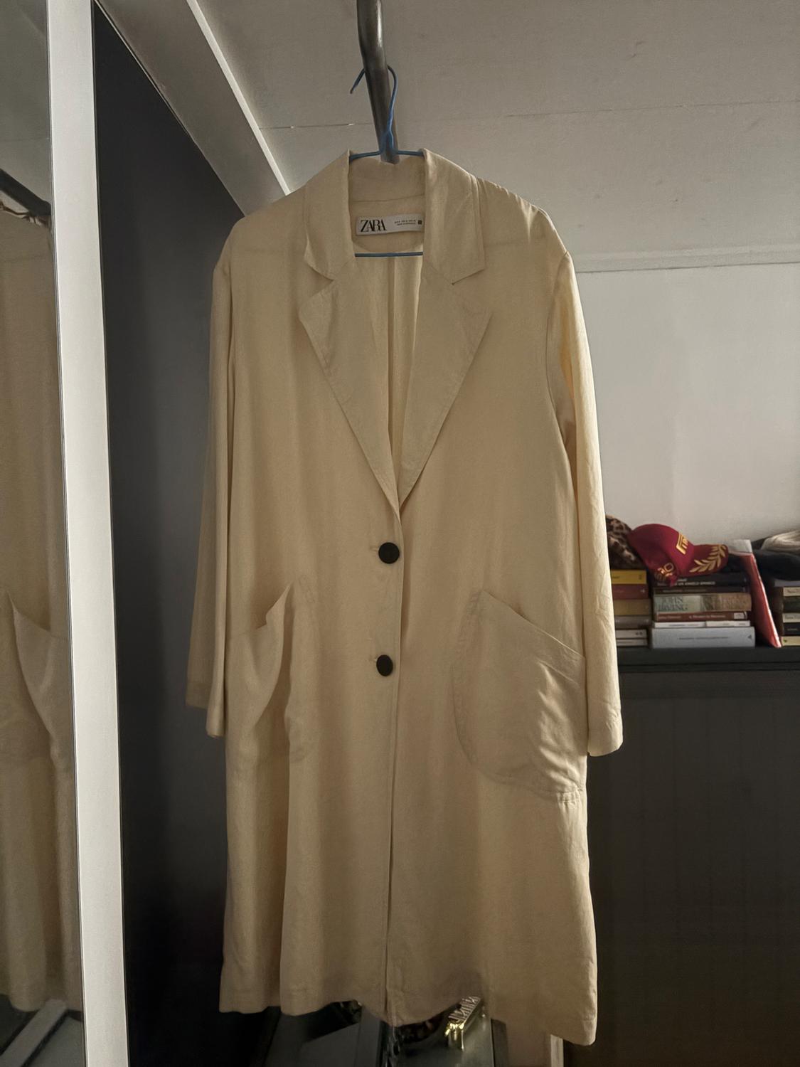 Beige coat hanging on a blue hanger, with two black buttons and large pockets. Background with stacked books and a red cap.