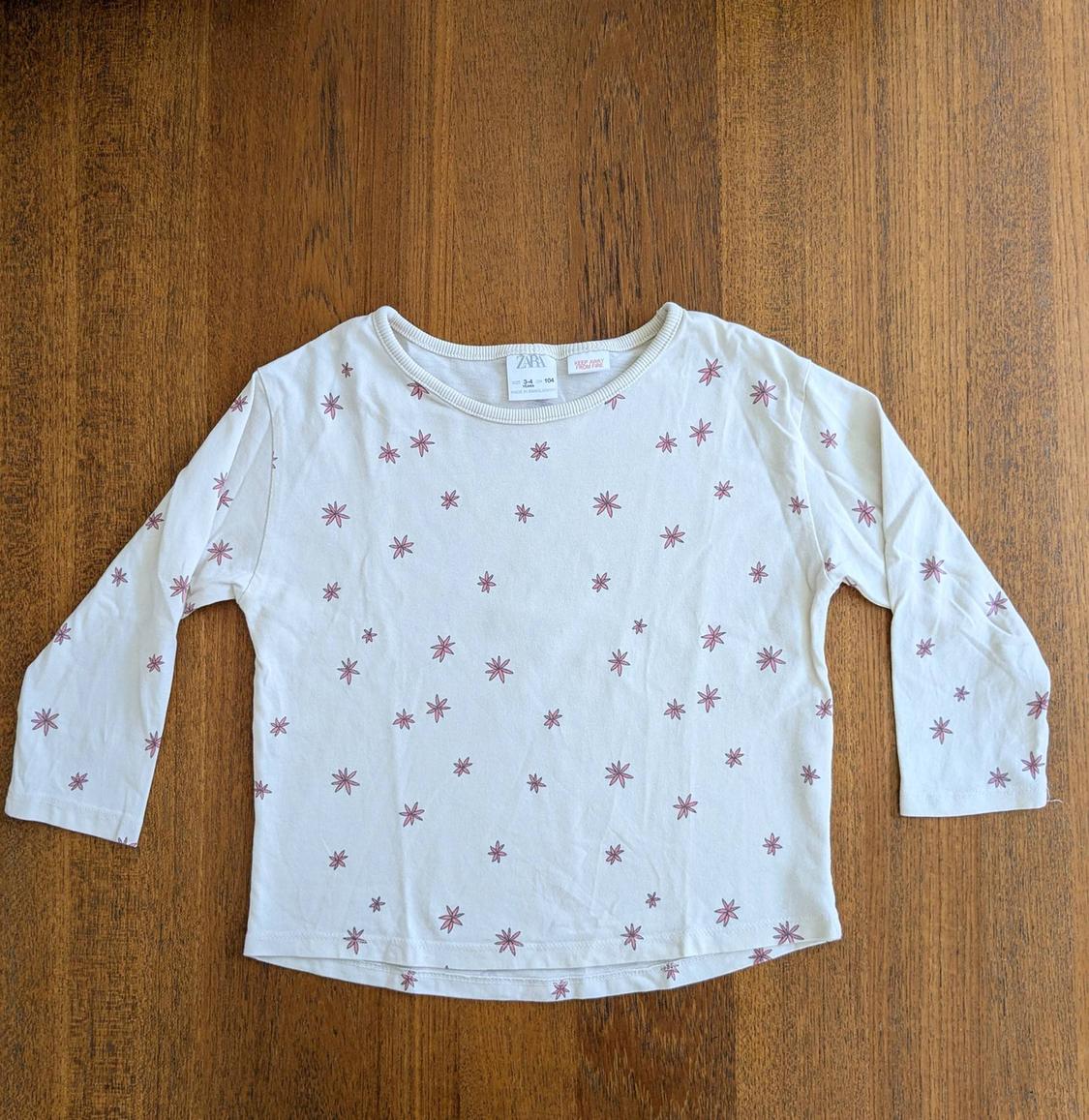 White long-sleeved t-shirt with a small pink floral print on a brown wood background. Visible label on the collar.