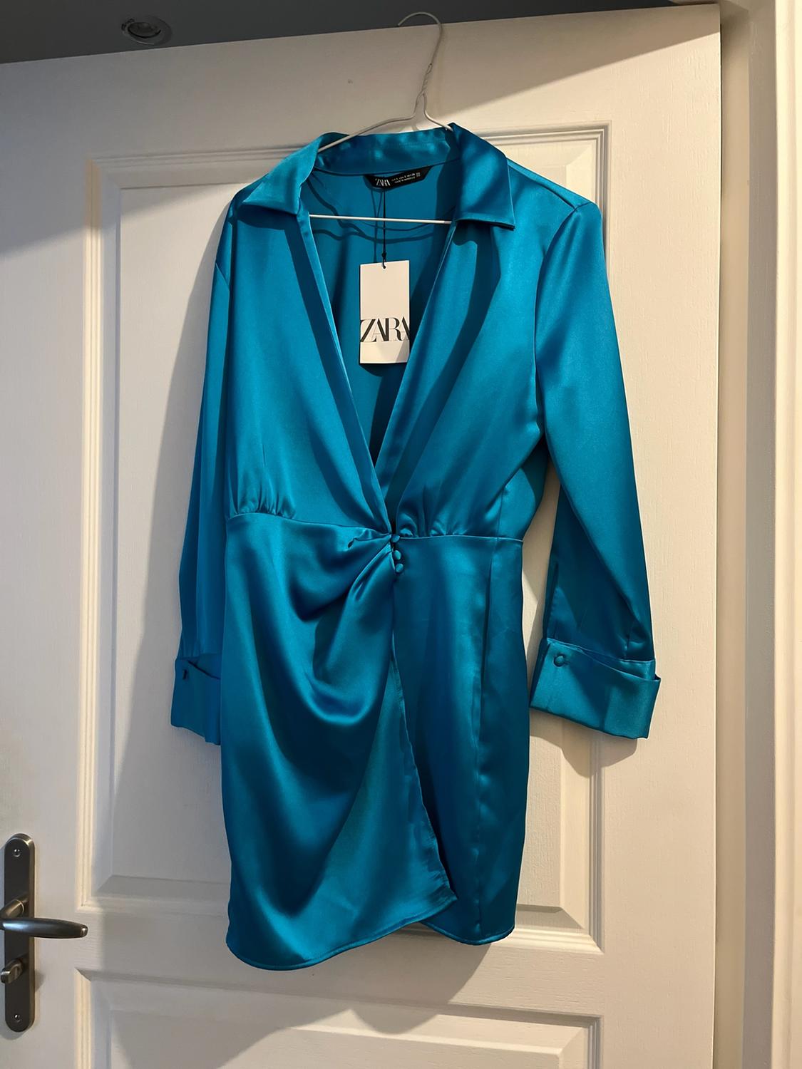 Short blue satin dress with a V-neck and ruched detail, hanging on a white door. Label with the text ZARA.