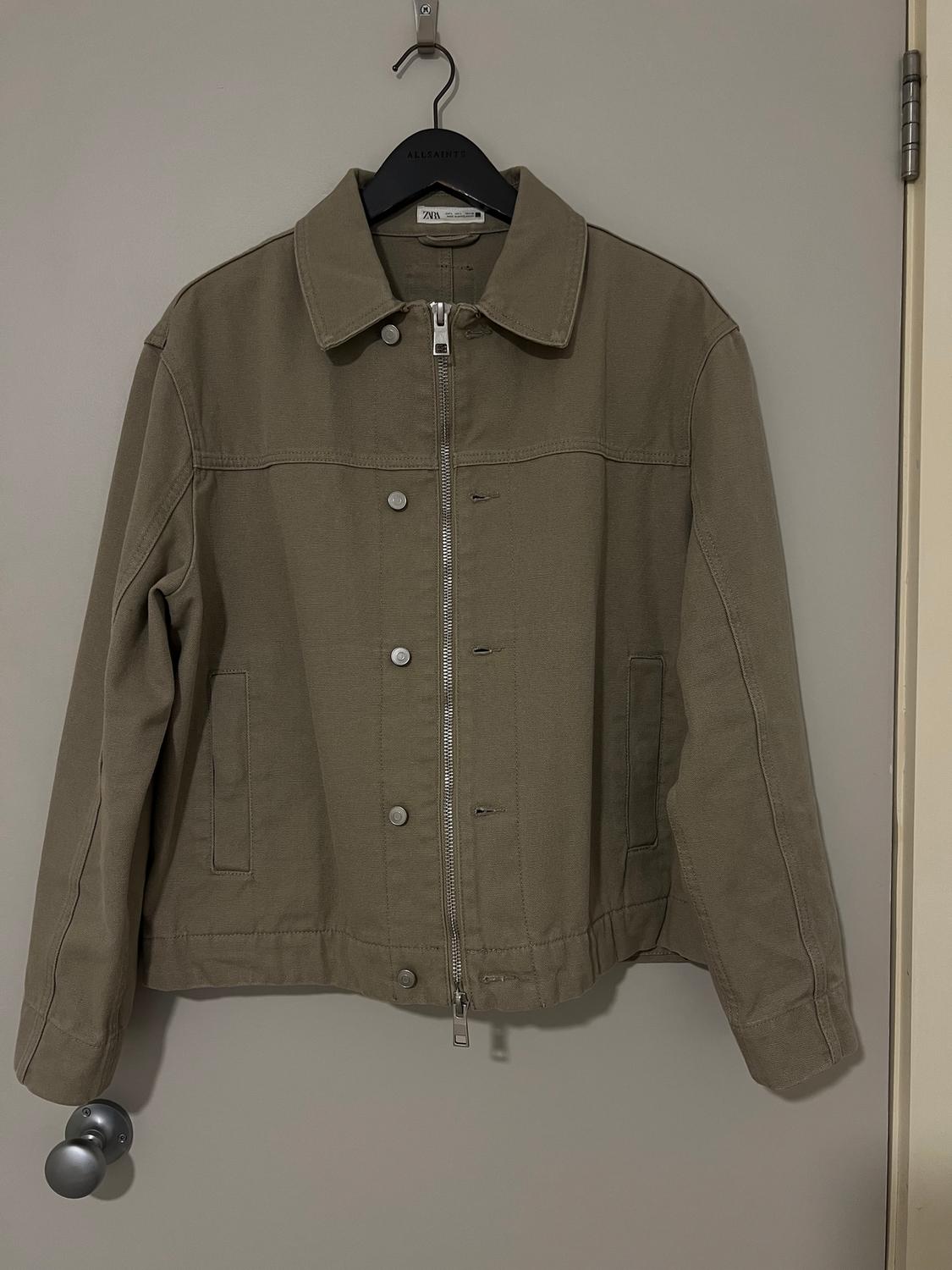 Khaki jacket with zip and metal buttons, hanging on a black AllSaints hanger over a grey door.
