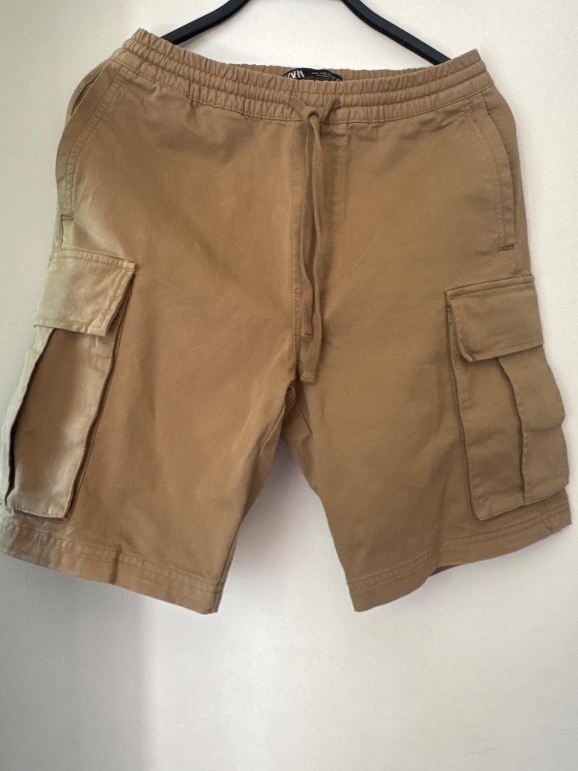 Beige cargo shorts with an elasticated waist, drawstring, and side pockets, hanging on a black hanger against a white wall.