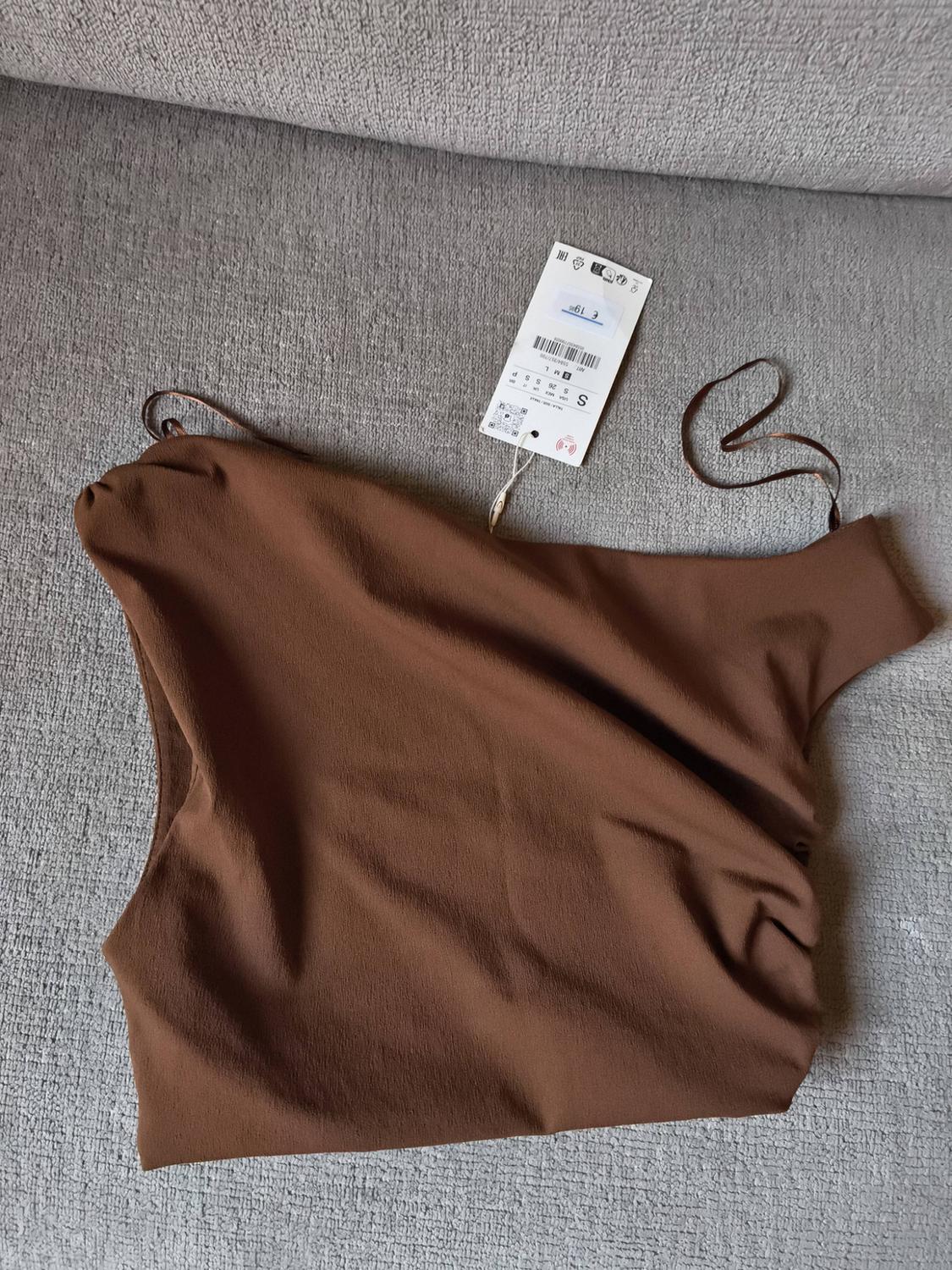Brown one-shoulder top on a grey sofa, with visible tag showing product details and size S.