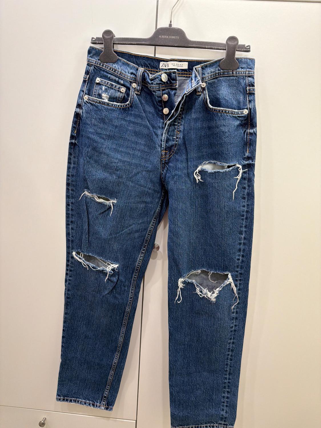 Distressed blue jeans with rips on the legs, hanging on a black hanger with an "Alberta Ferretti" tag.
