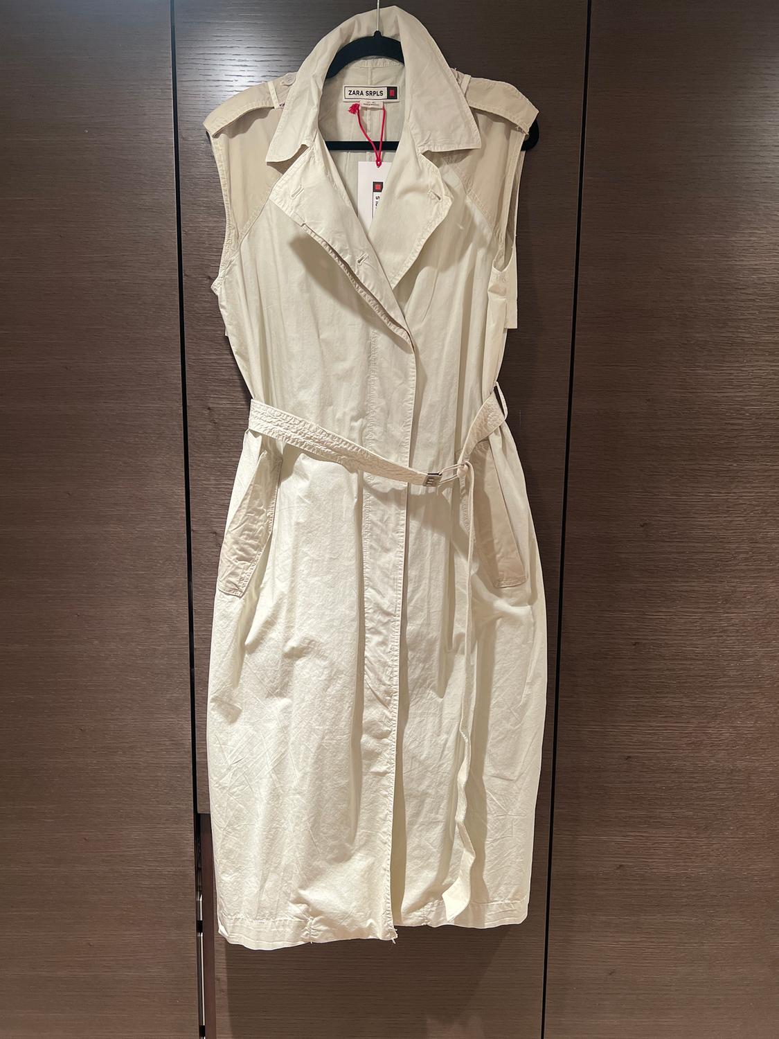 Cream sleeveless trench coat hanging on a hanger, with a belt and wide lapels. Visible label: "ZARA SRPLS". Dark wooden background.