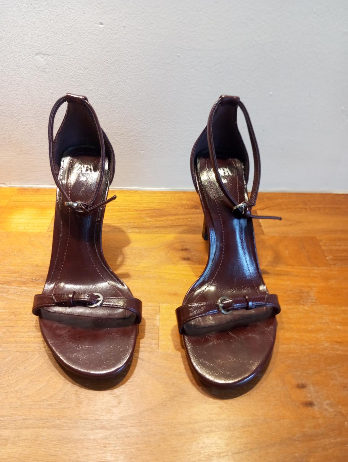 Dark brown high-heeled sandals with thin straps and buckles, on a light wooden surface.