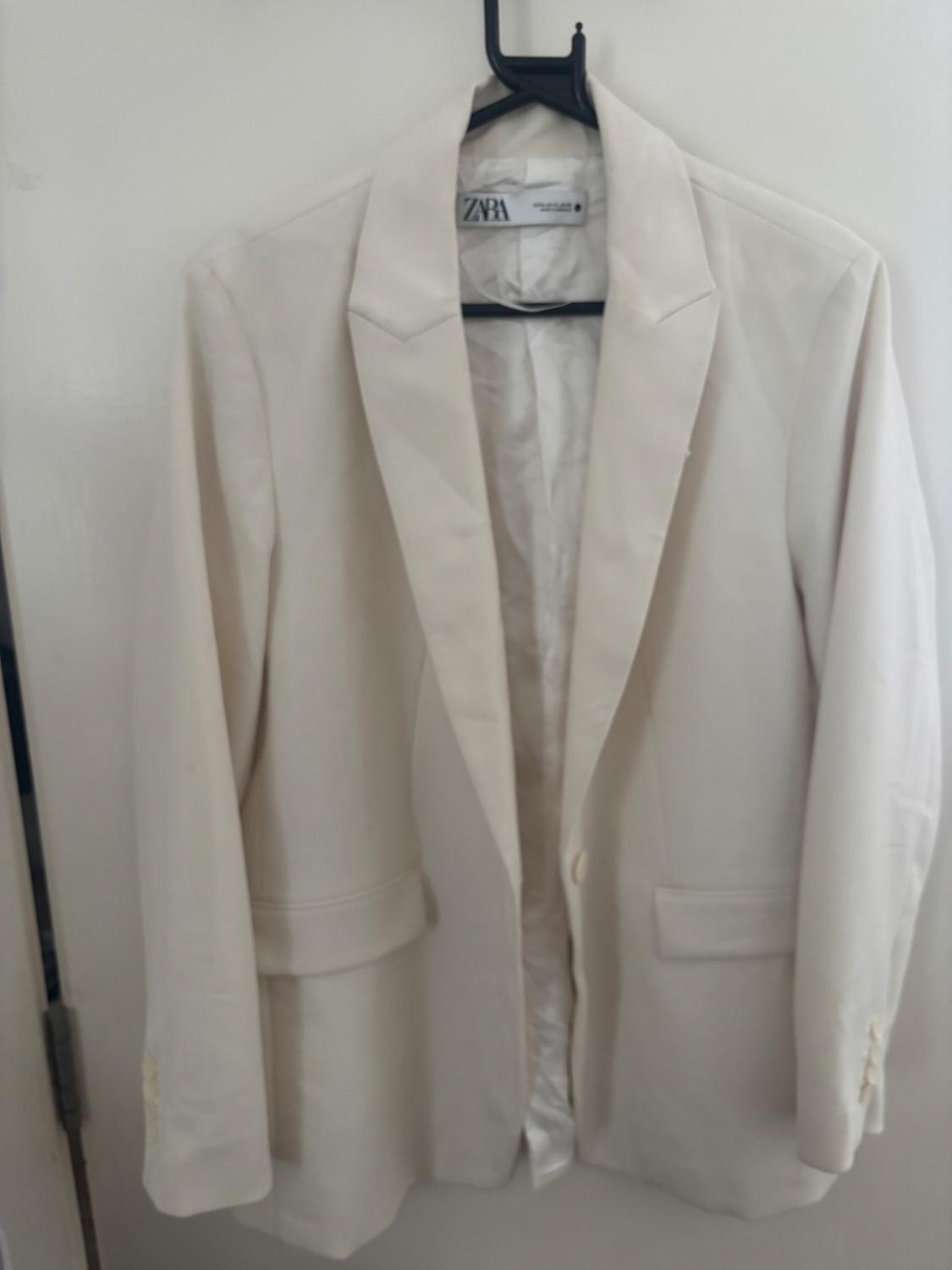 White jacket hanging on a black hanger, with a visible "ZARA" tag. White background.
