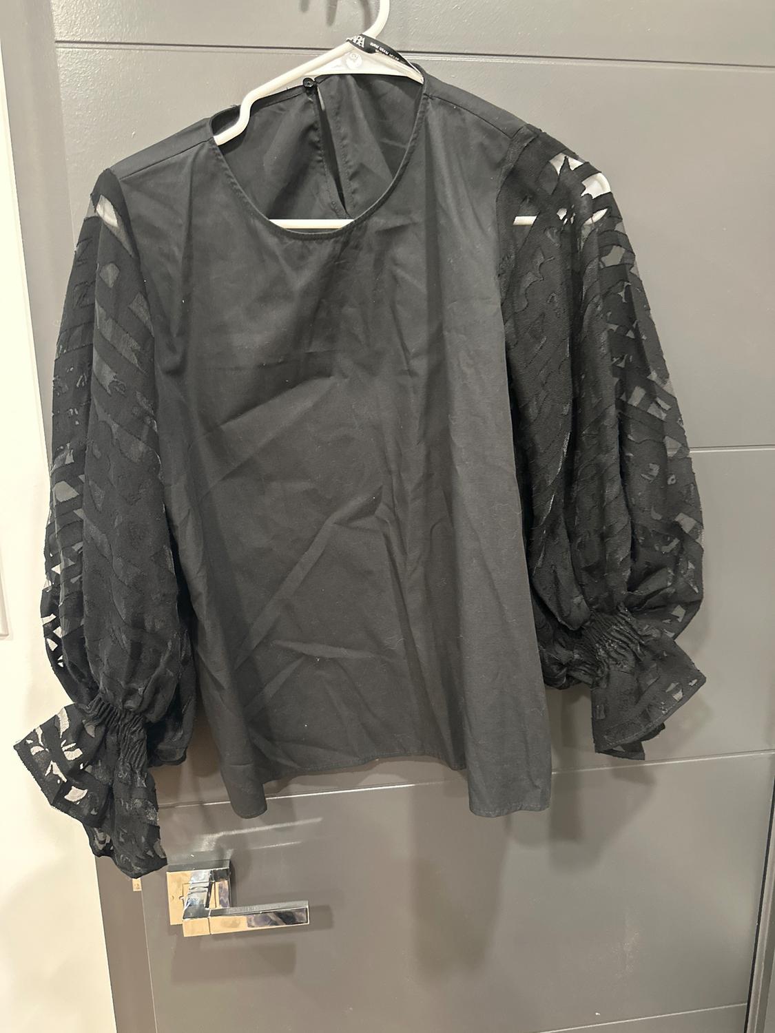 Black blouse with long, sheer sleeves hanging on a white hanger over a gray door.