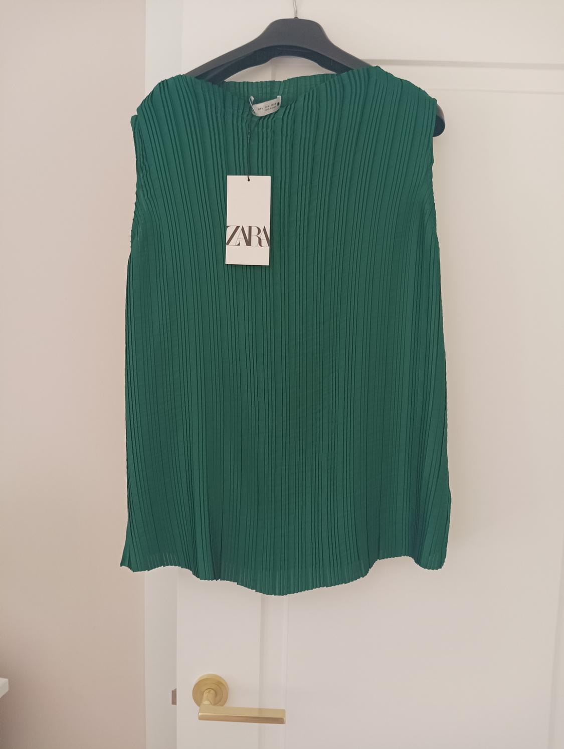 Pleated green dress hanging on a black hanger, with a white label that says "ZARA", on a white door with a gold handle.