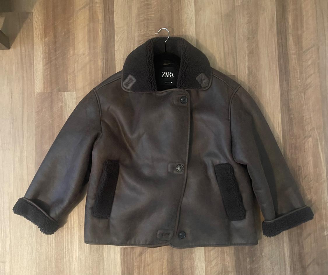 Brown faux leather jacket with black lining and buttons, hanging on a hanger over a wooden floor. Visible text: "ZARA".