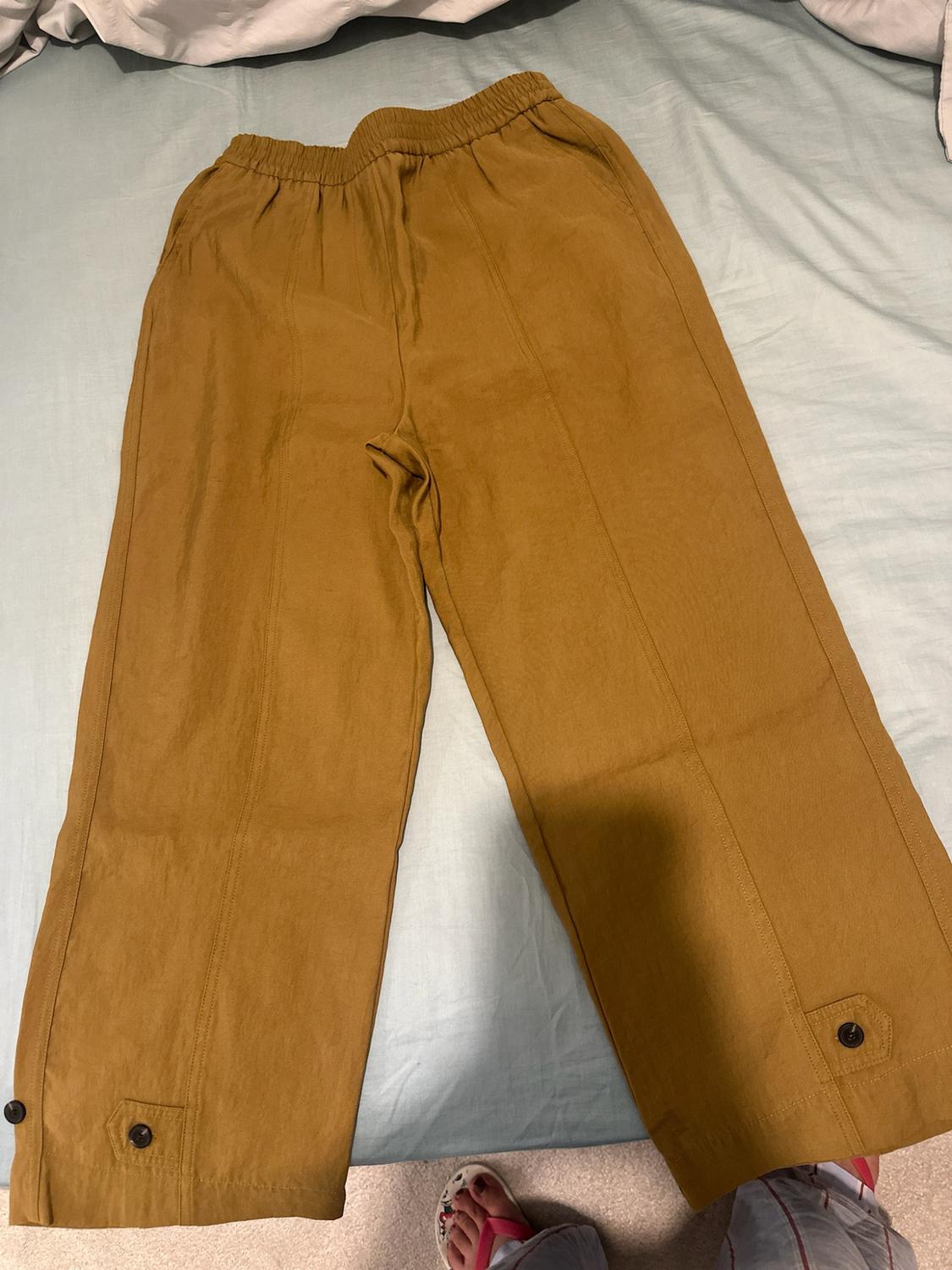 Mustard-coloured wide-leg trousers with an elasticated waist, front seams and buttons at the hem, laid out on a bed with light blue sheets.