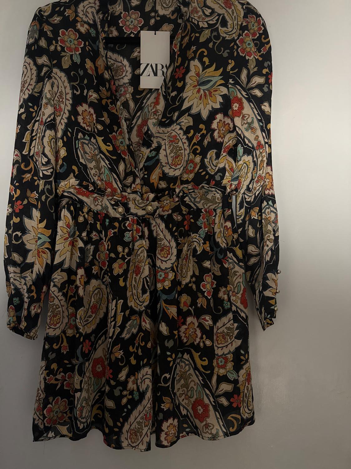 Black dress with colorful floral print and belt, hanging on a hanger. Visible tag with text "ZARA".