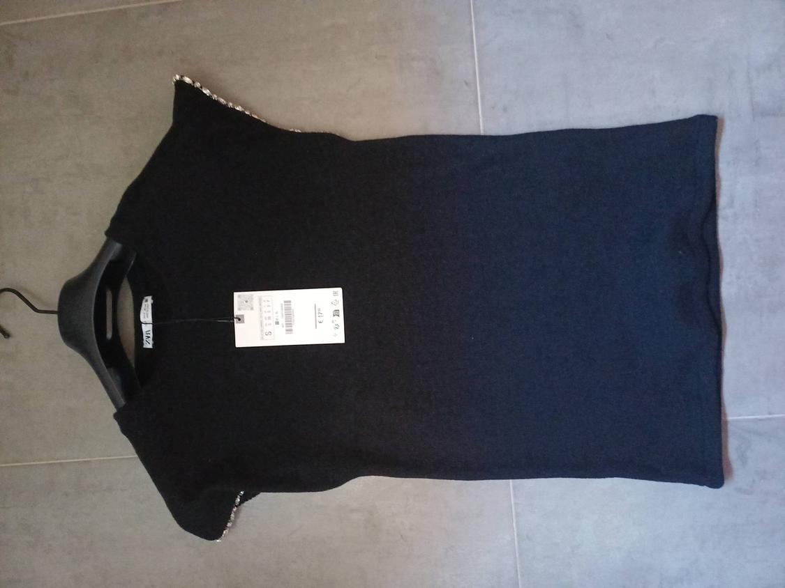 Black short-sleeved top with rhinestones on the shoulders, on a hanger on a grey tiled floor. Visible text: ZARA, S, € 17.