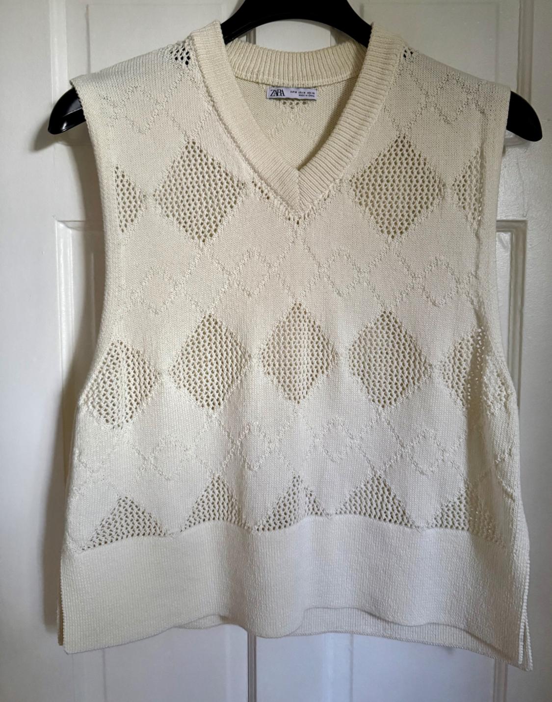 Cream-colored sleeveless knit vest with a V-neck and a diamond cut-out pattern, hanging on a hanger over a white door.