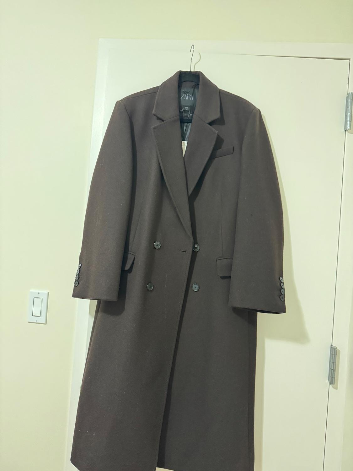 Brown wool coat hanging on a white door, with wide lapels and black buttons. Visible tag on the inside.