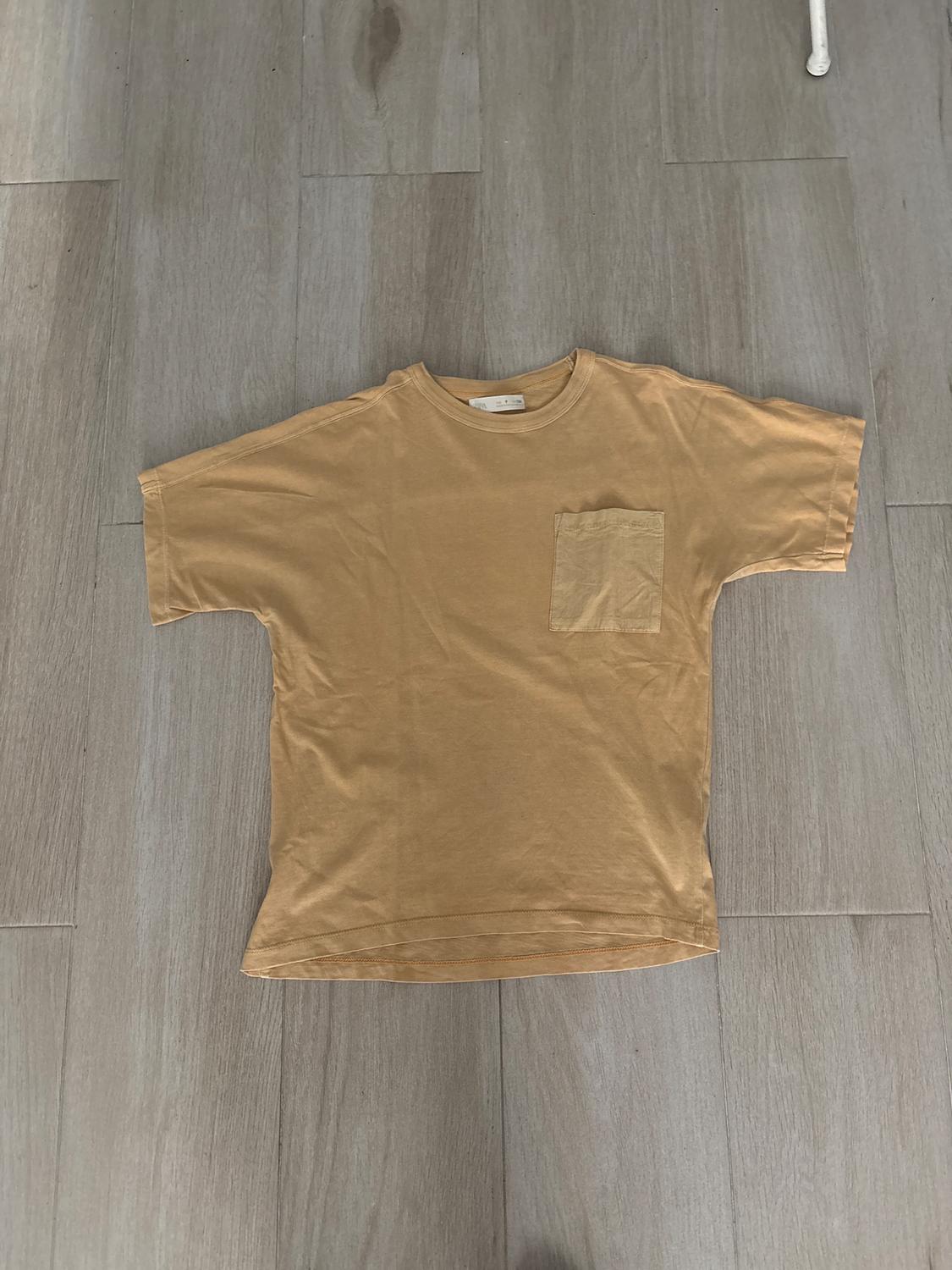 Light brown t-shirt with a chest pocket, placed on a grey wooden floor.