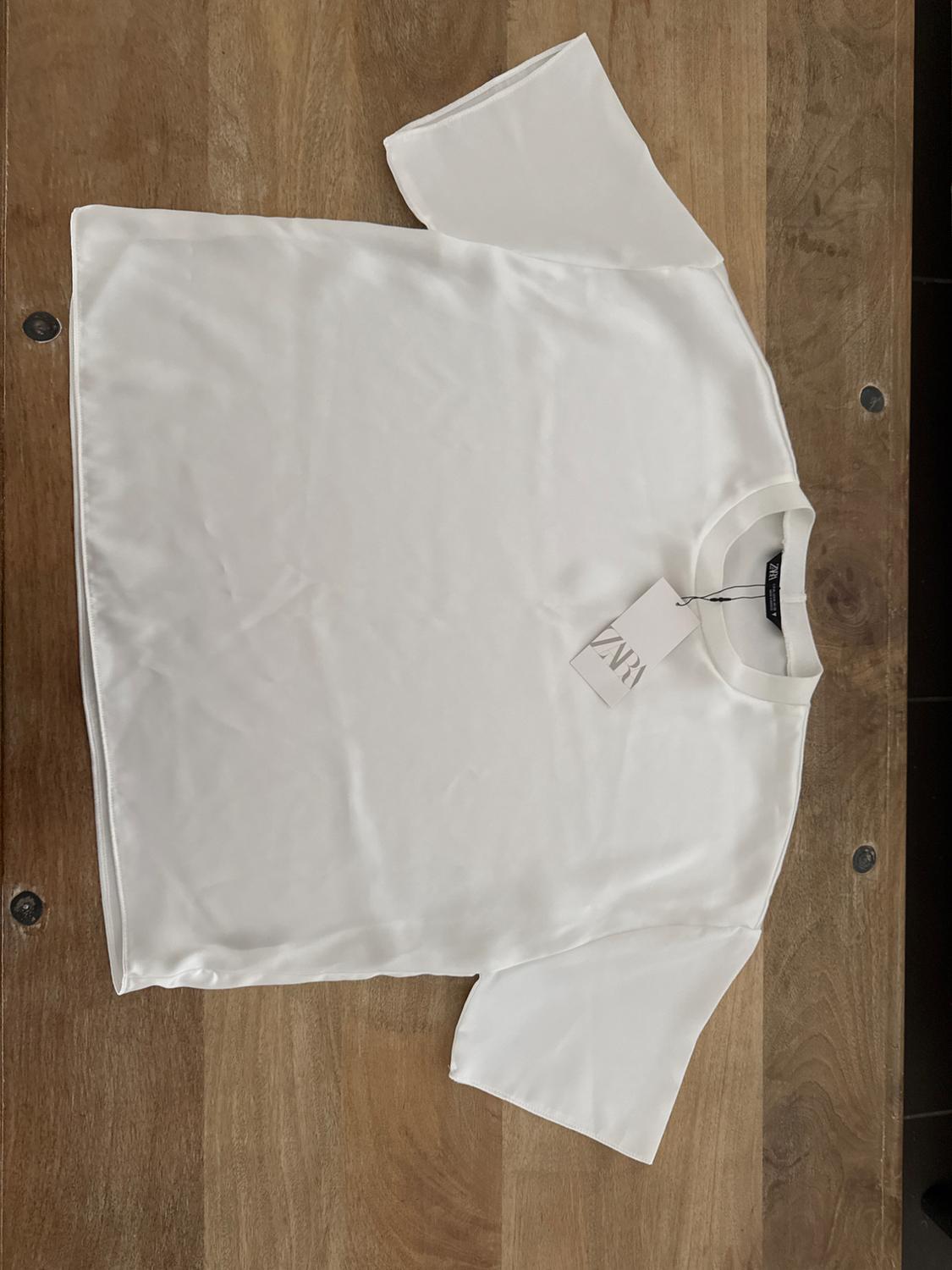 White, short-sleeved T-shirt with a round neck, visible Zara label, on a light wood surface.