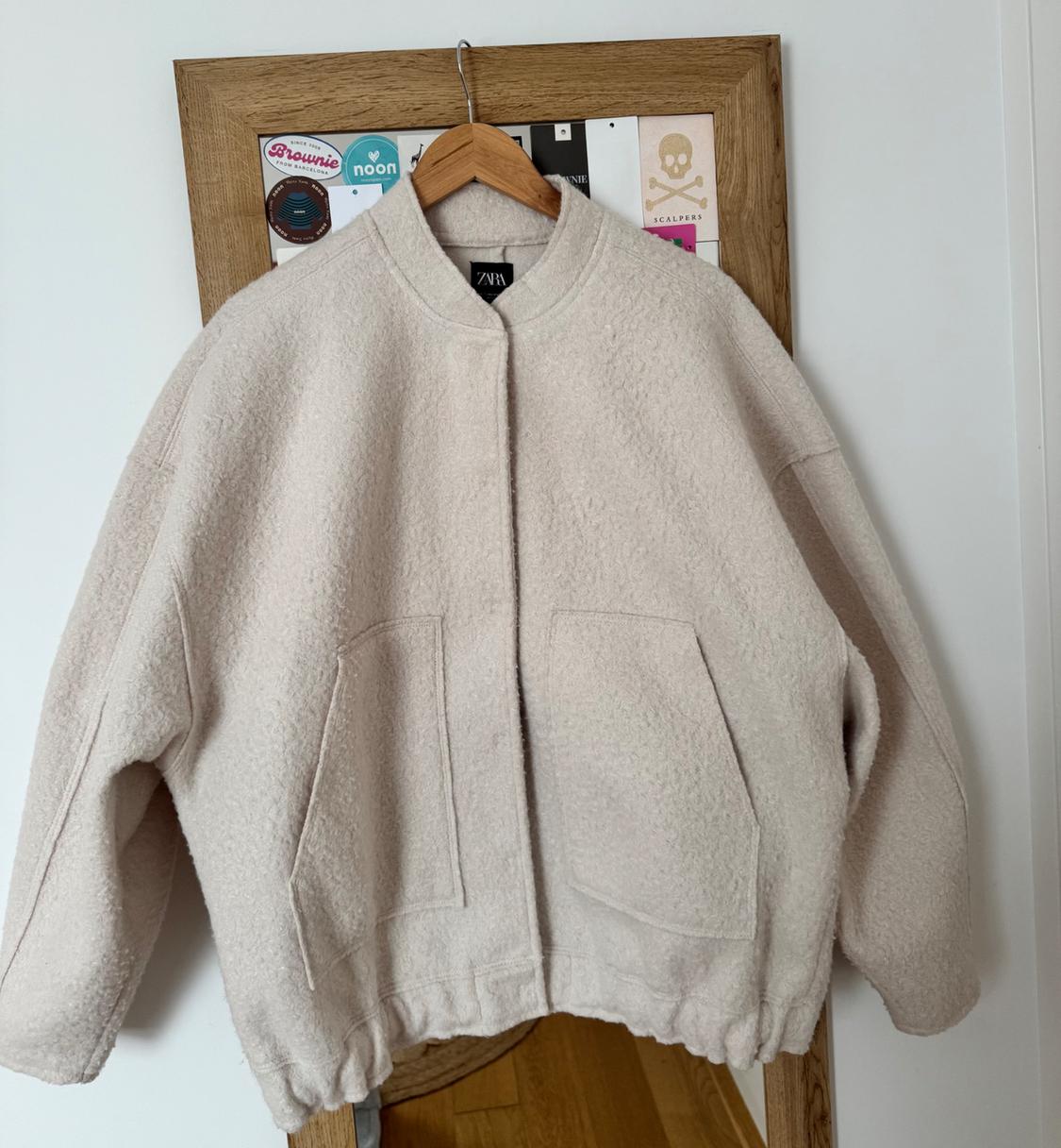 Beige wool jacket hanging on a hanger in front of a mirror with cards and stickers. Visible brand: ZARA.