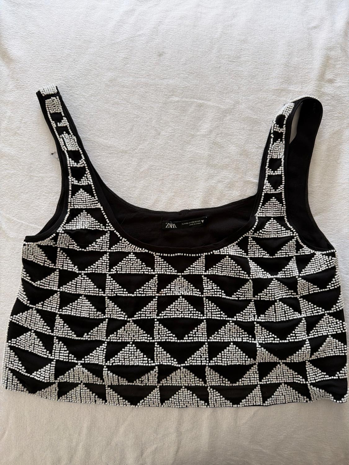 Black sleeveless top with white beading forming triangles, on a white textured fabric. Label with ZARA text.