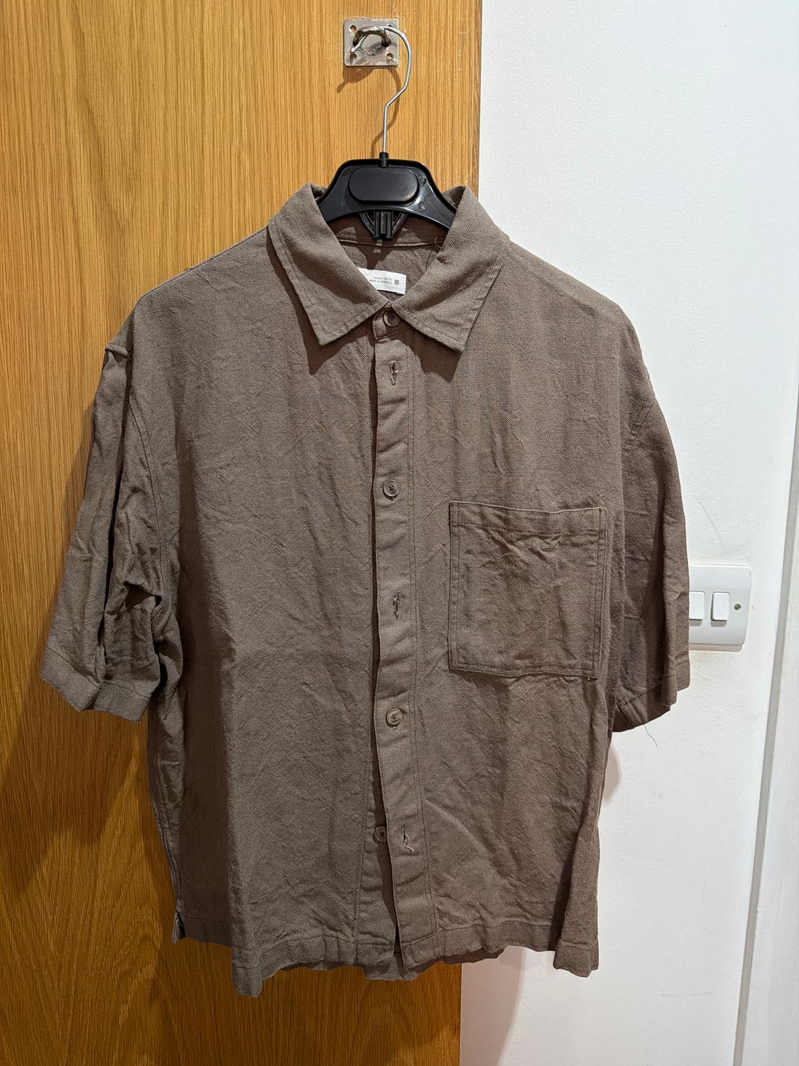 Short-sleeved brown shirt hanging on a black hanger over a light wooden door. Chest pocket.