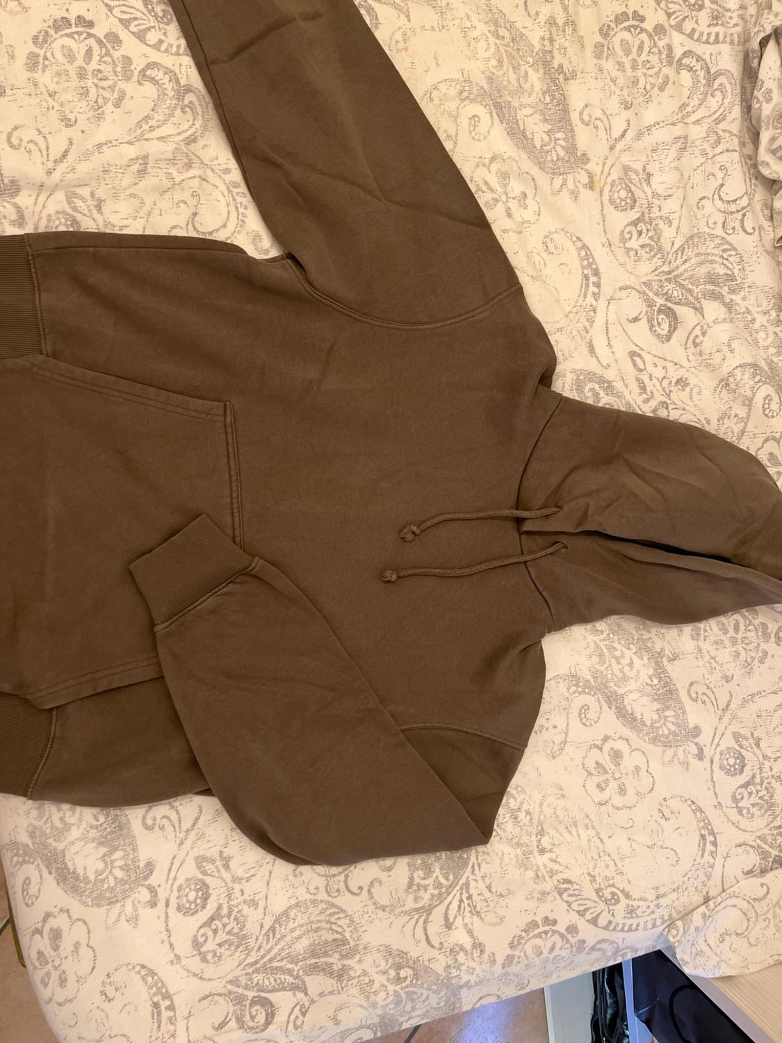 Brown hooded sweatshirt with drawstring and front pocket, on a light floral-print sheet in a relaxed setting.