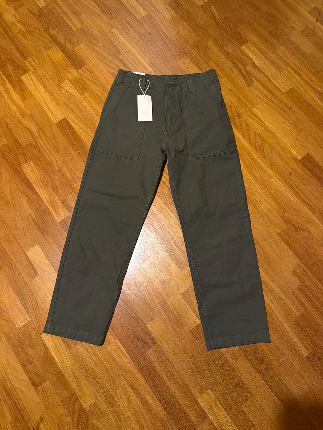 Dark green trousers with large pockets, white hang tag, on light wooden flooring.