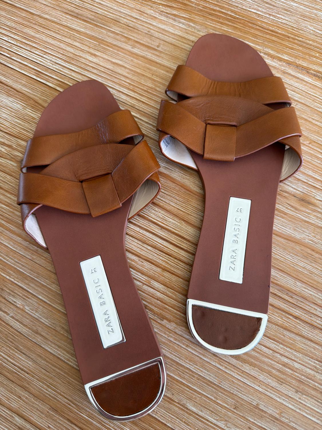 Brown flat sandals with interwoven straps on a wooden background. Visible label: "ZARA BASIC".