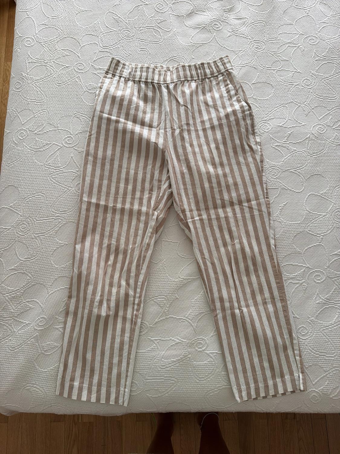 Beige and white vertical striped trousers with an elasticated waistband, on a white bedspread with floral embossing. Some feet are peeking out at the bottom.