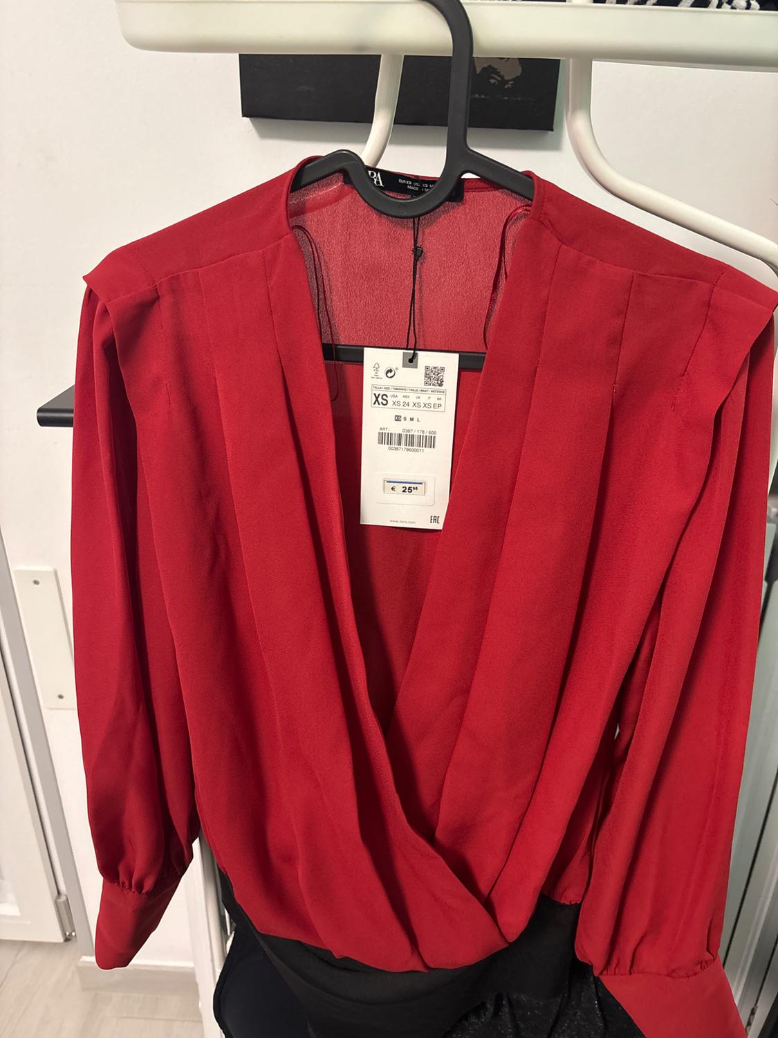 Long-sleeved red blouse hanging on a hanger, with an XS size tag and a price of €25. Black and white background.