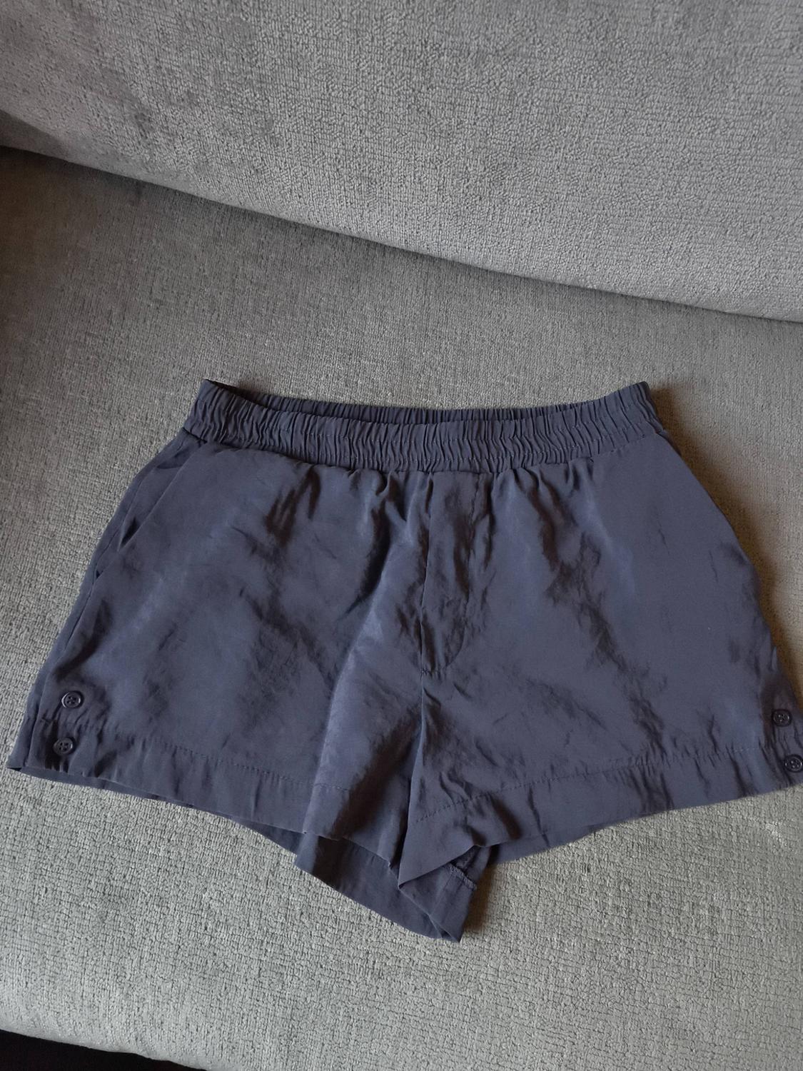 Grey shorts with an elasticated waist and buttons on the sides, placed on a grey sofa.
