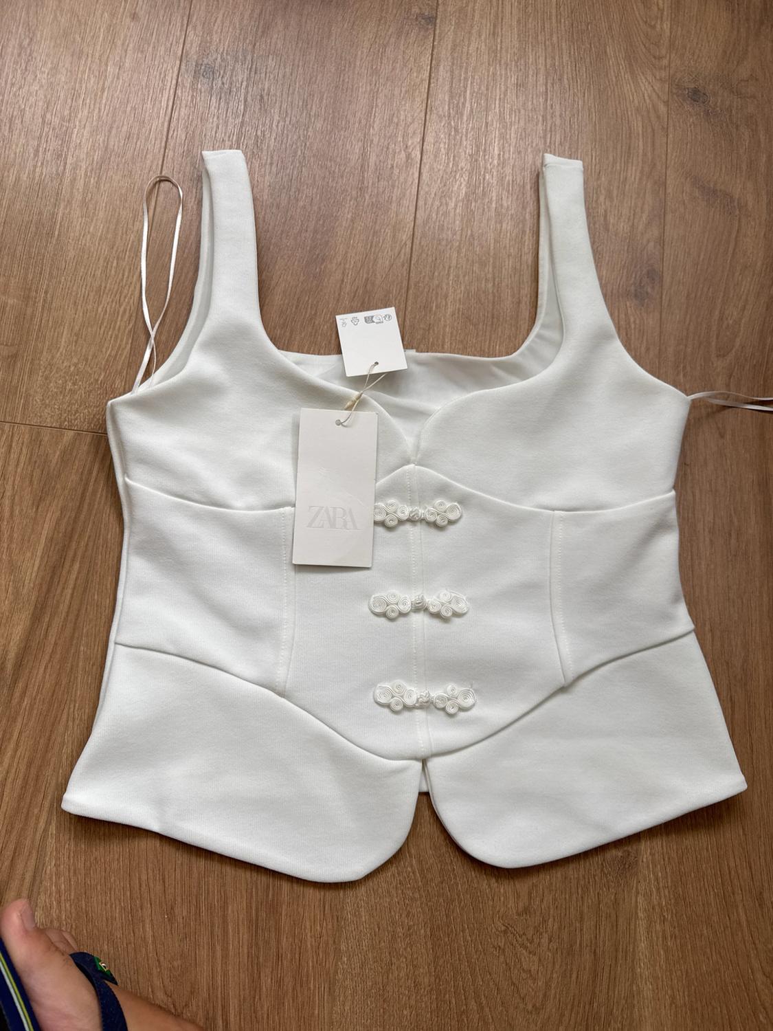 White sleeveless top with decorative front knot fastenings, on a wooden floor. Visible label bearing the text ZARA.