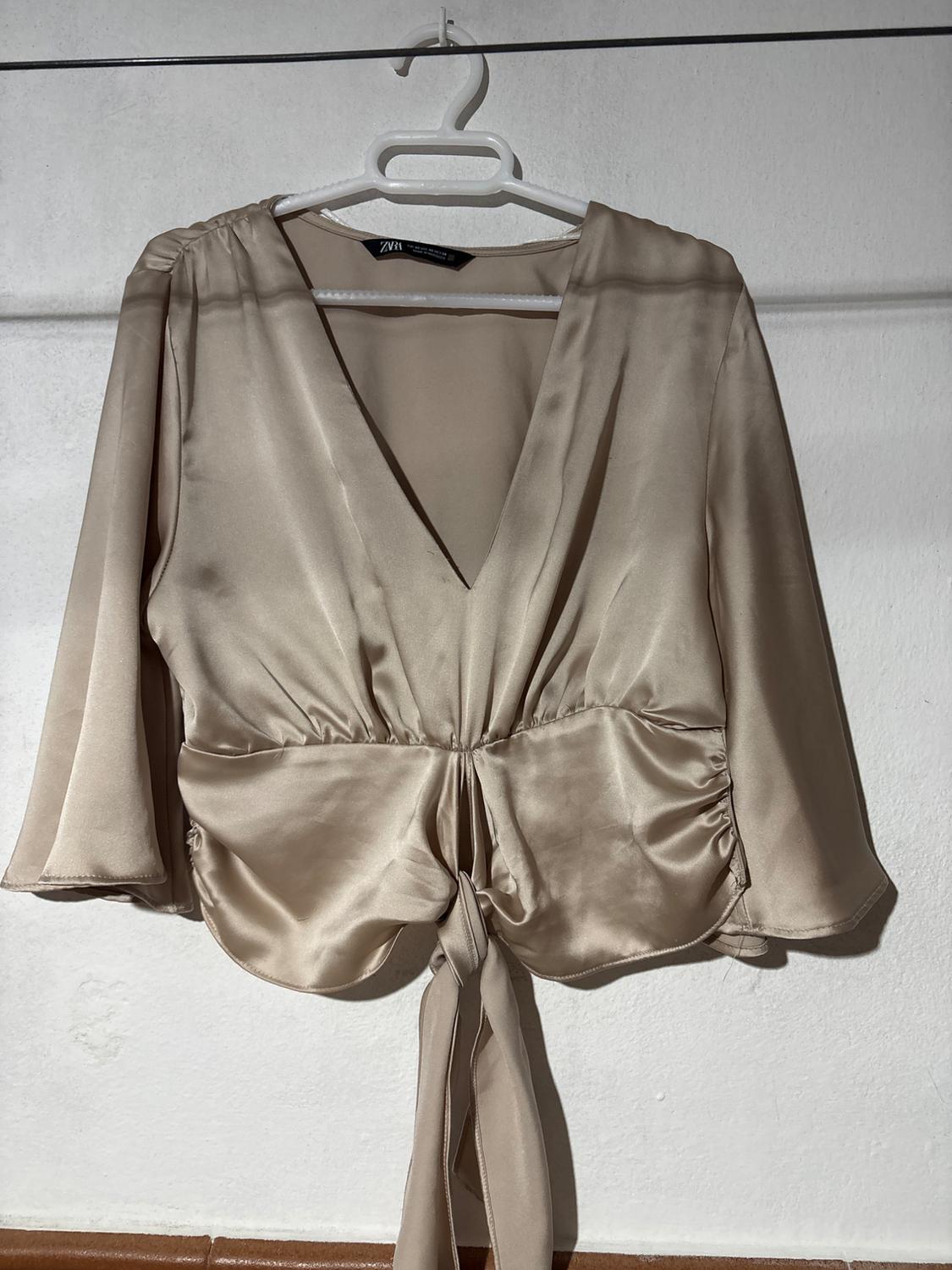 Beige satin blouse with V-neck and long sleeves, hanging on a white hanger against a white background.