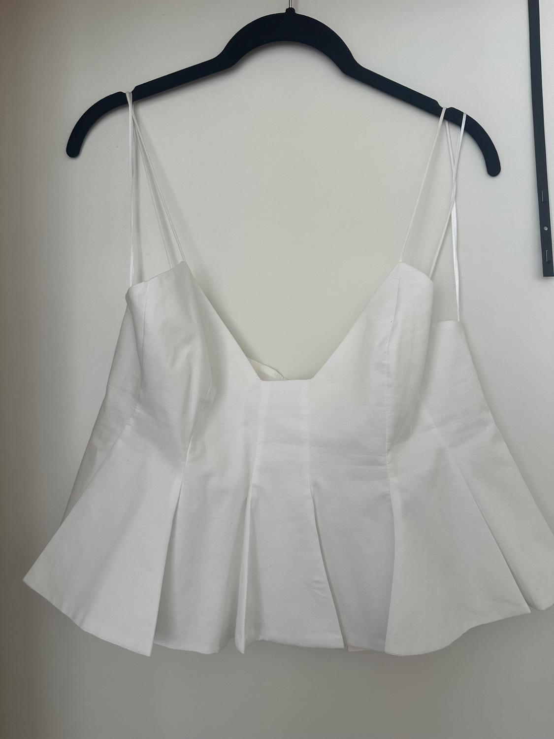 White top with double spaghetti straps, V-neck and pleated hem, hanging on a black hanger against a white wall. Minimalist style.