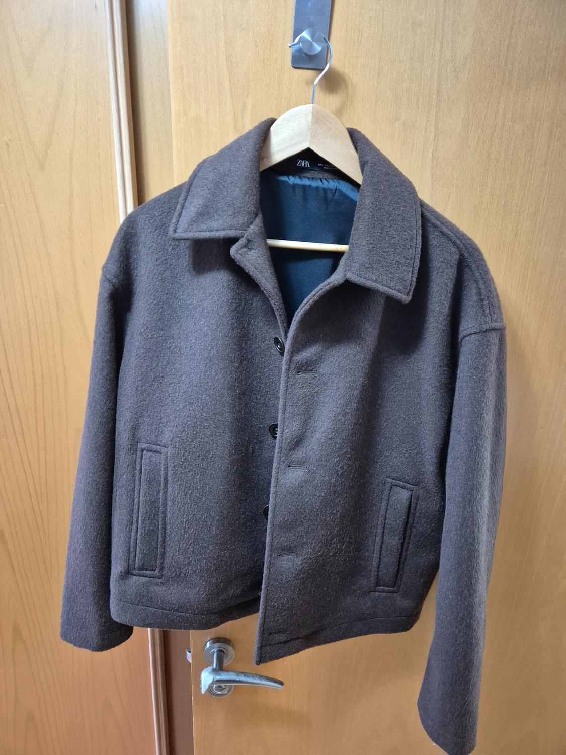 Brown wool jacket hanging on a wooden hanger over a light wooden door. Label visible on the collar.