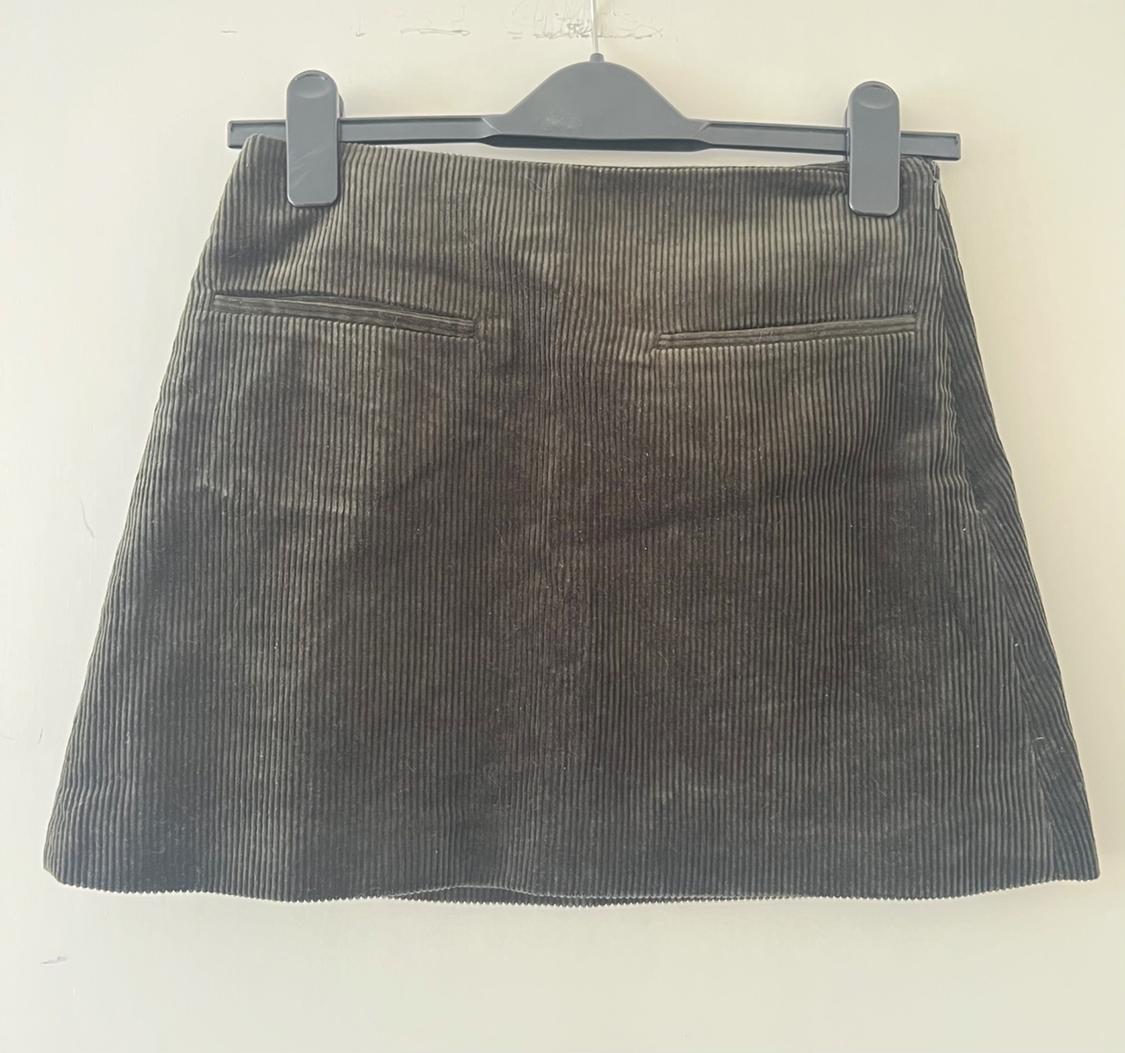 Dark green corduroy mini skirt with back pockets, hanging from a black clip hanger against a light-coloured wall background.