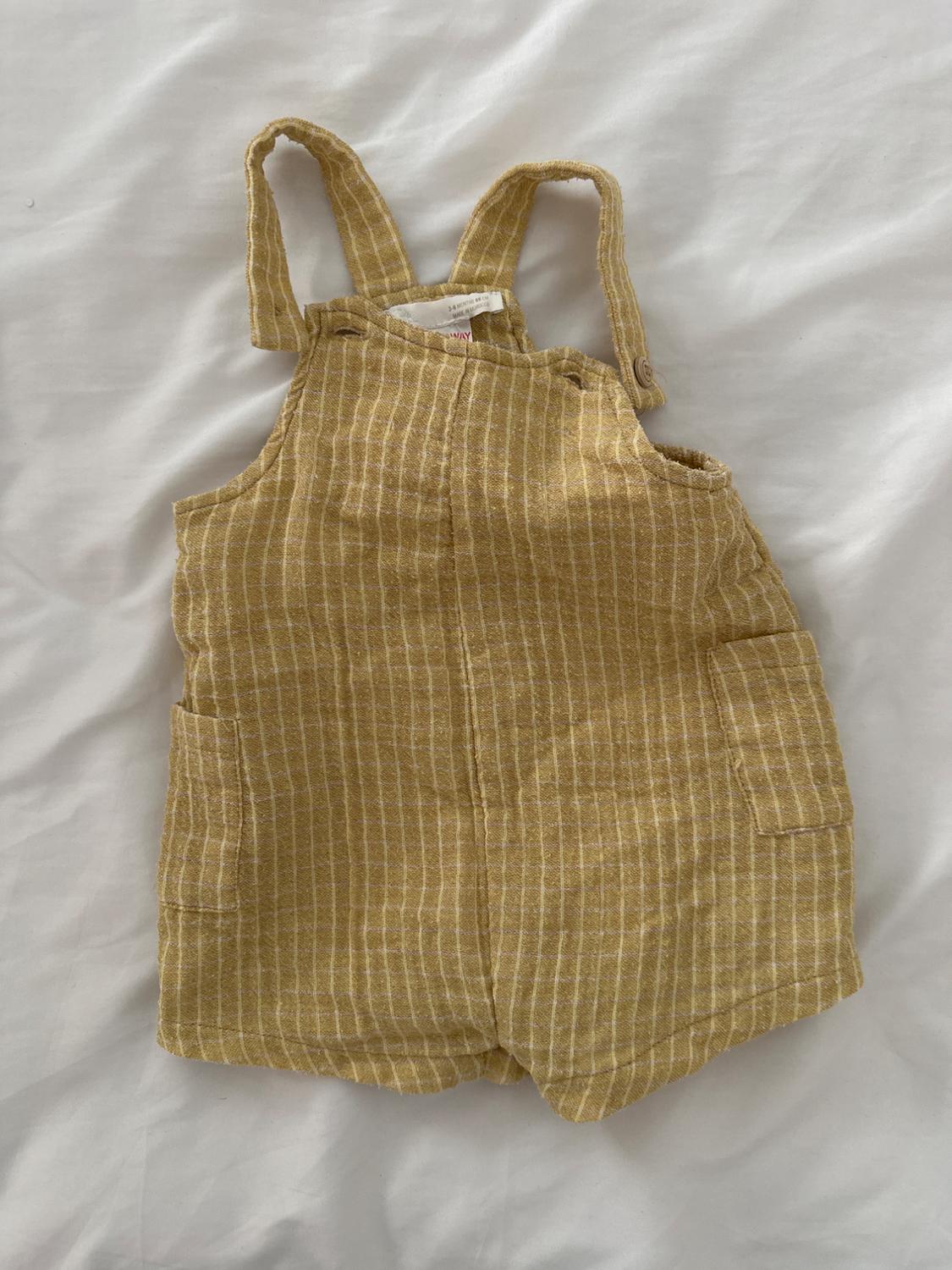Children's yellow fabric dungarees with white stripes, straps and front pockets, on a white background. Label visible on the inside.