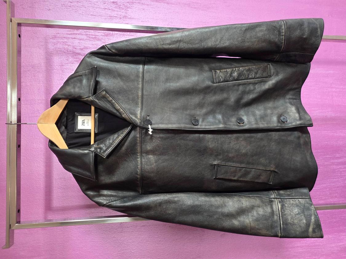 Black jacket hanging on a wooden hanger against a pink background. The inside label shows the brand "Zara".
