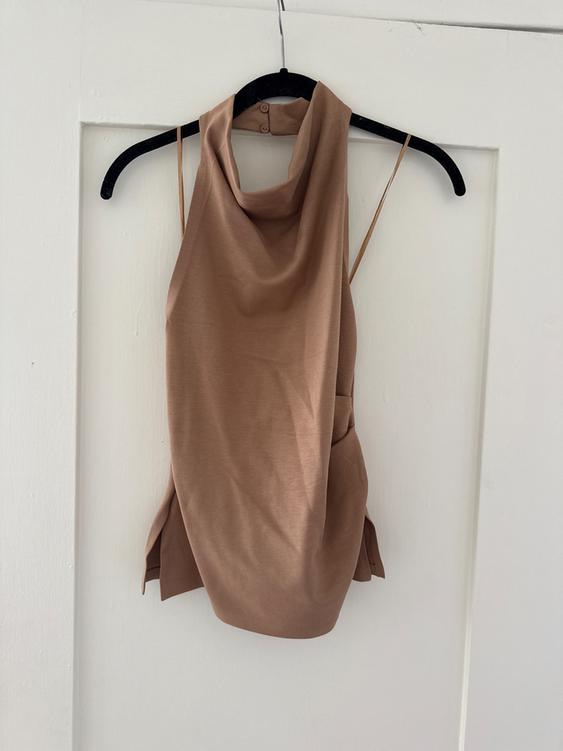 Beige sleeveless top hanging on a black hanger over a white door. Design with a draped neckline and thin straps.
