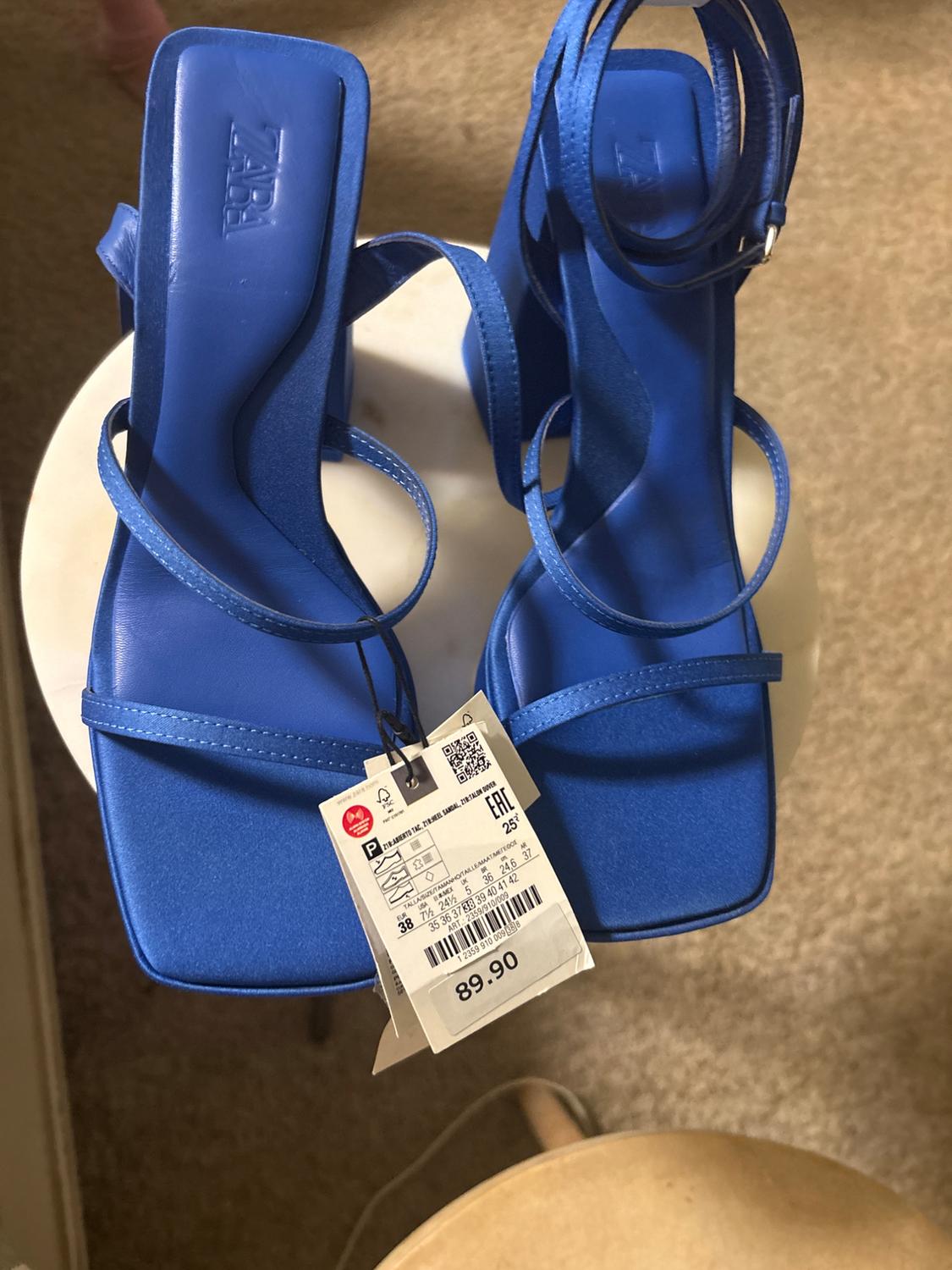 Blue high-heeled sandals with thin straps, price tag of 89.90 on a white table, beige carpet background.