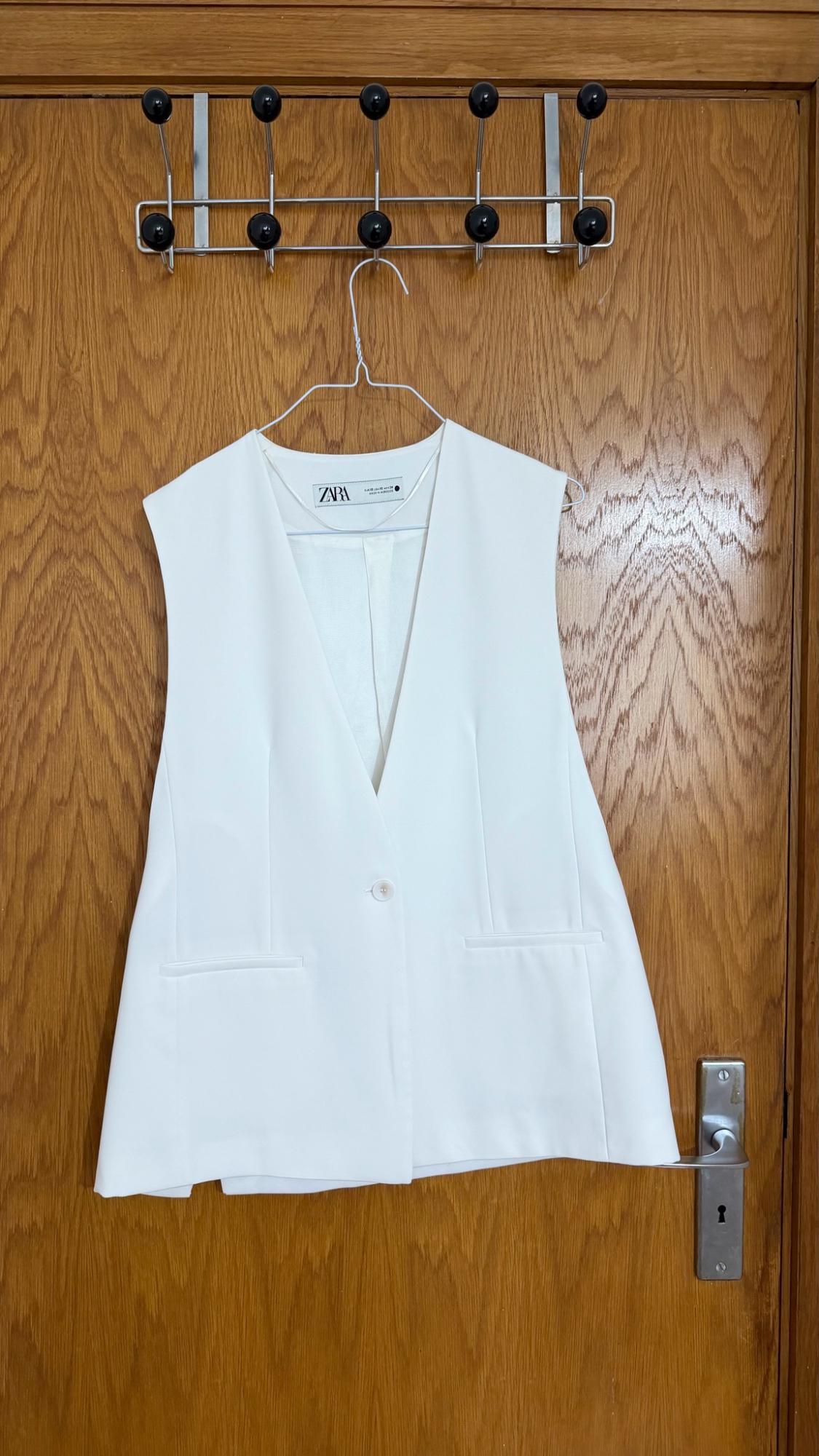 White waistcoat with a V-neck, one button and pockets, hanging on a wooden door. Label bearing the text ZARA.