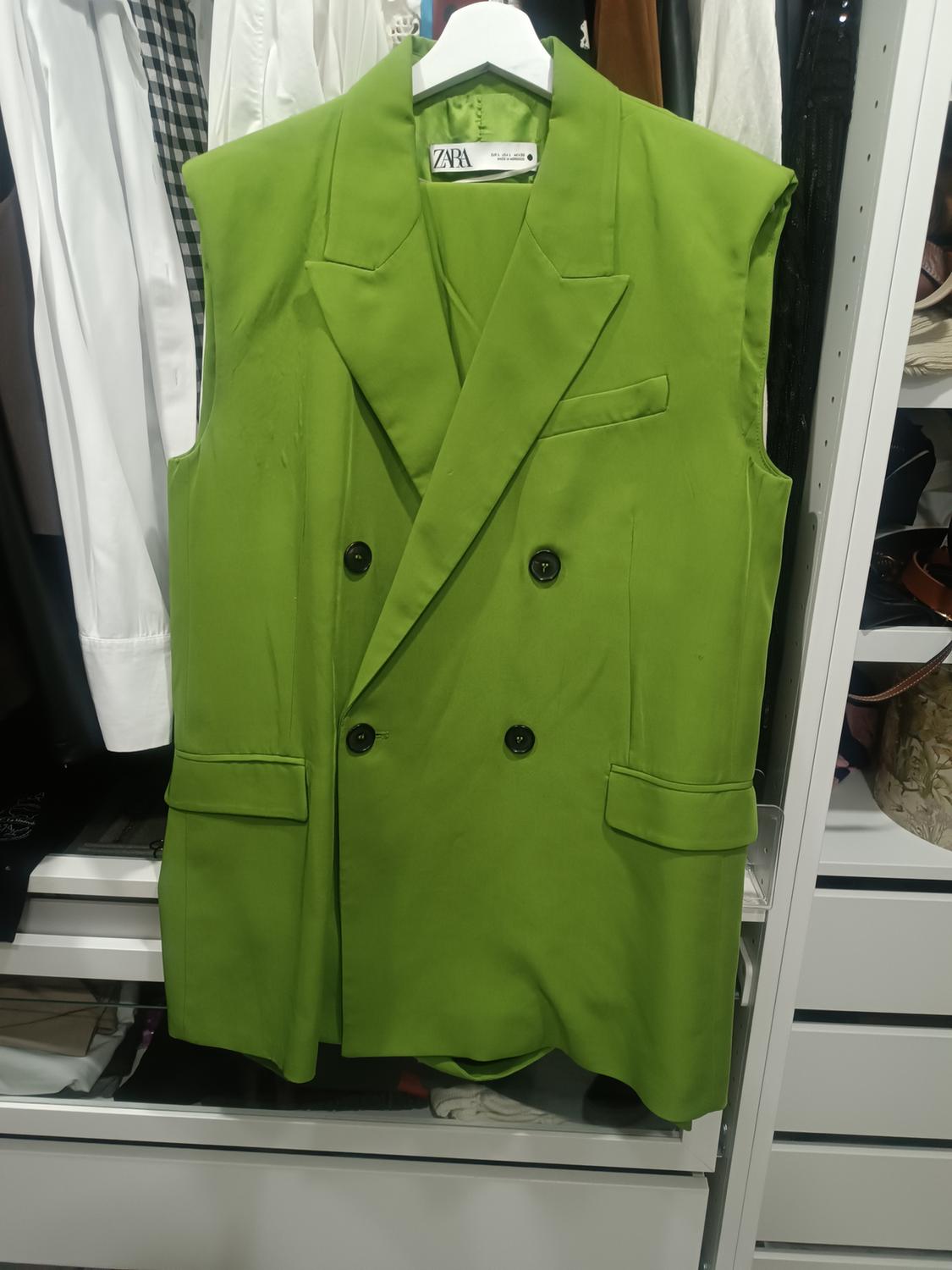 Green sleeveless double-breasted waistcoat hanging in a wardrobe, with visible Zara label. Background with assorted clothes.