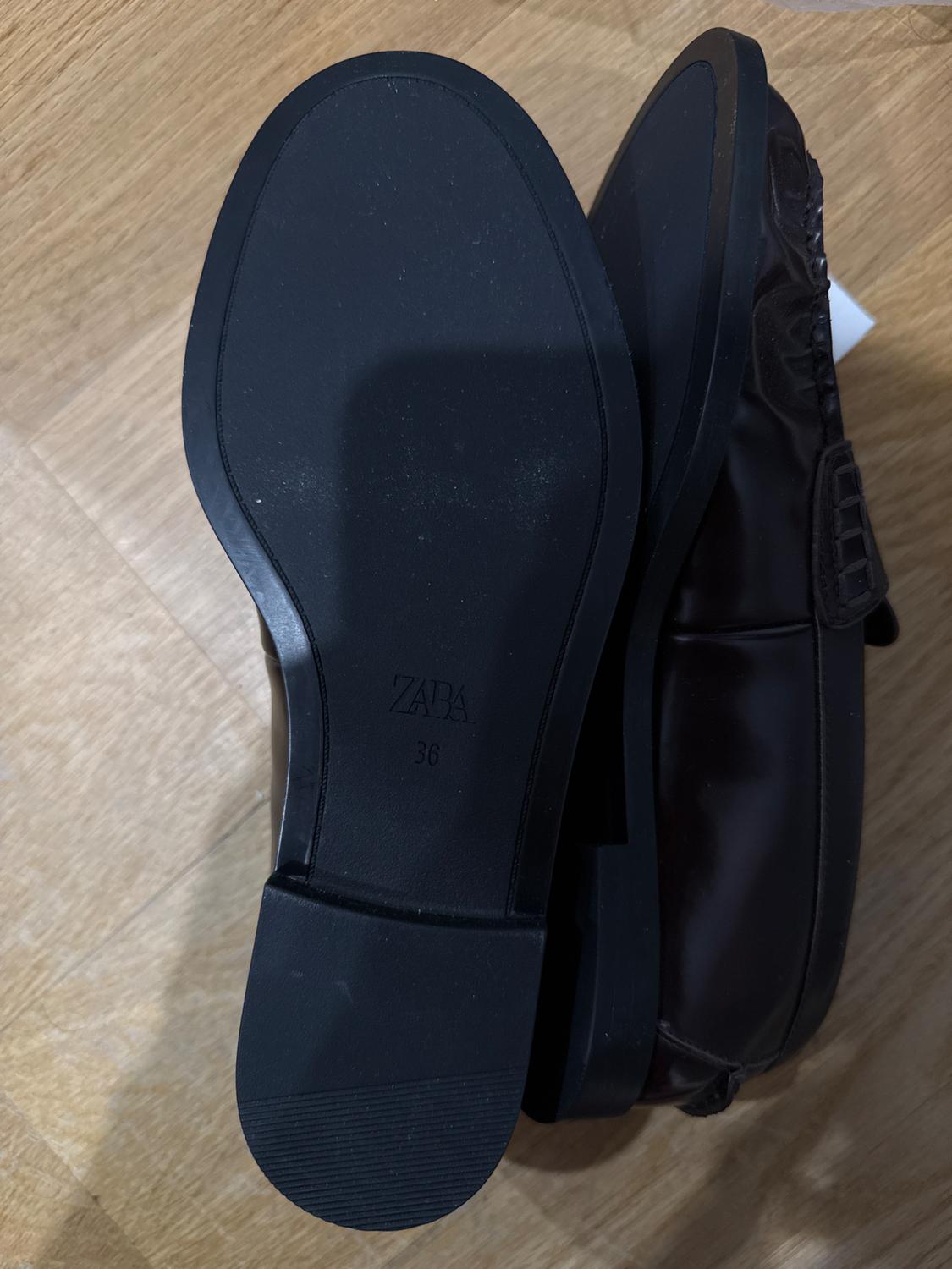 Pair of dark loafers on a wooden floor. One shows its black sole with the text ZARA 36, the other rests on its side.