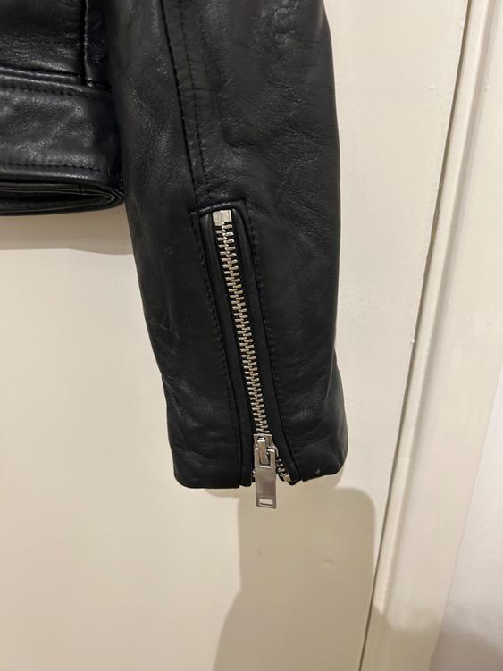 Black leather jacket sleeve with visible silver zip, hanging over a white background.