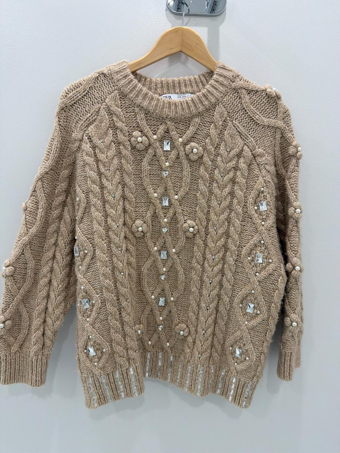 Beige cable-knit sweater on a wooden hanger. Decorated with pearls, sparkling appliqués, and flowers. Tag with ZARA text.